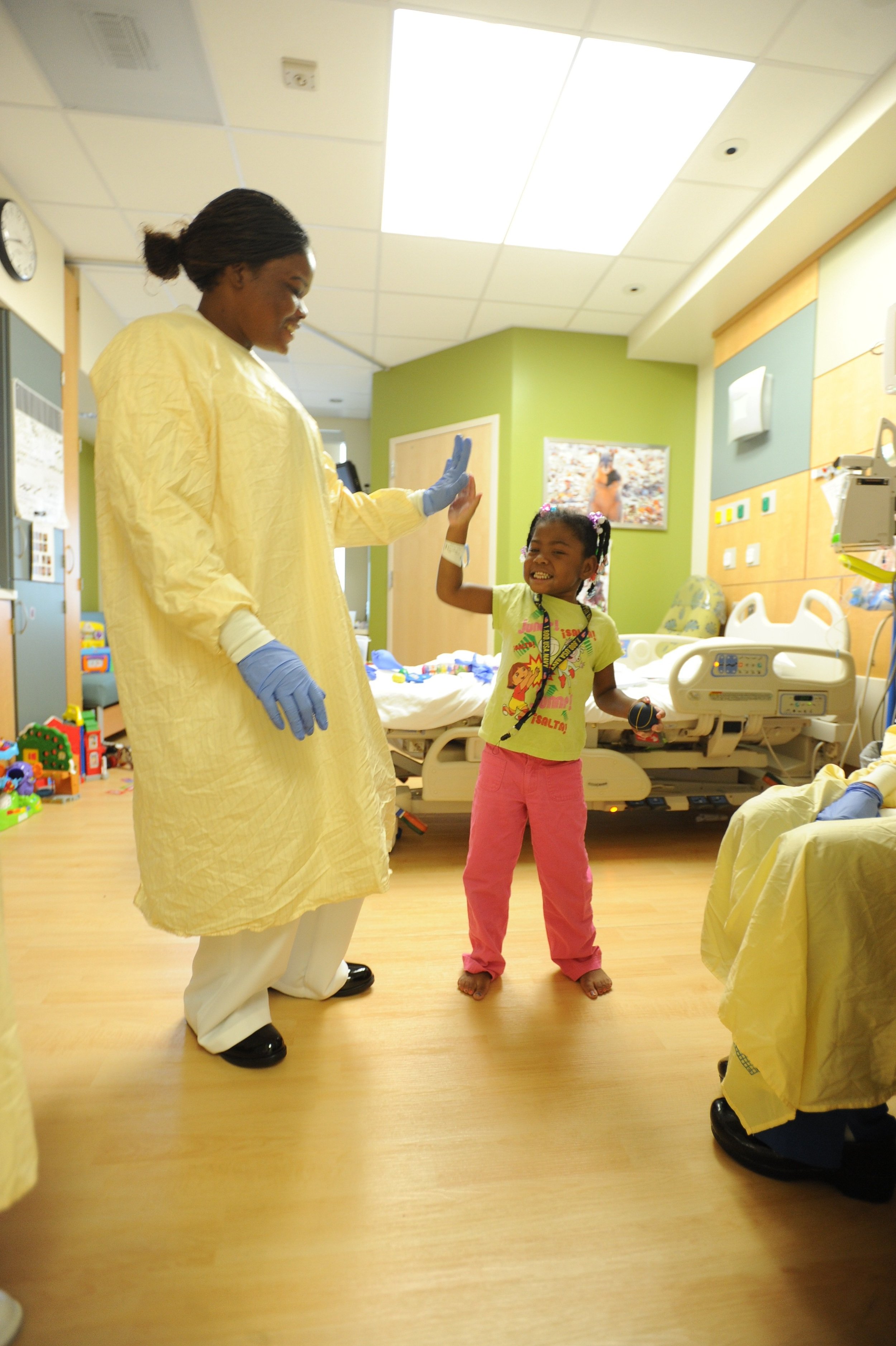 A nurse or healthcare worker giving a high-five to a young girl in a hospital room. The girl is smiling and dressed in pink pants and a colorful shirt. The room has yellow and green walls, and medical equipment is visible, including hospital beds and toys.