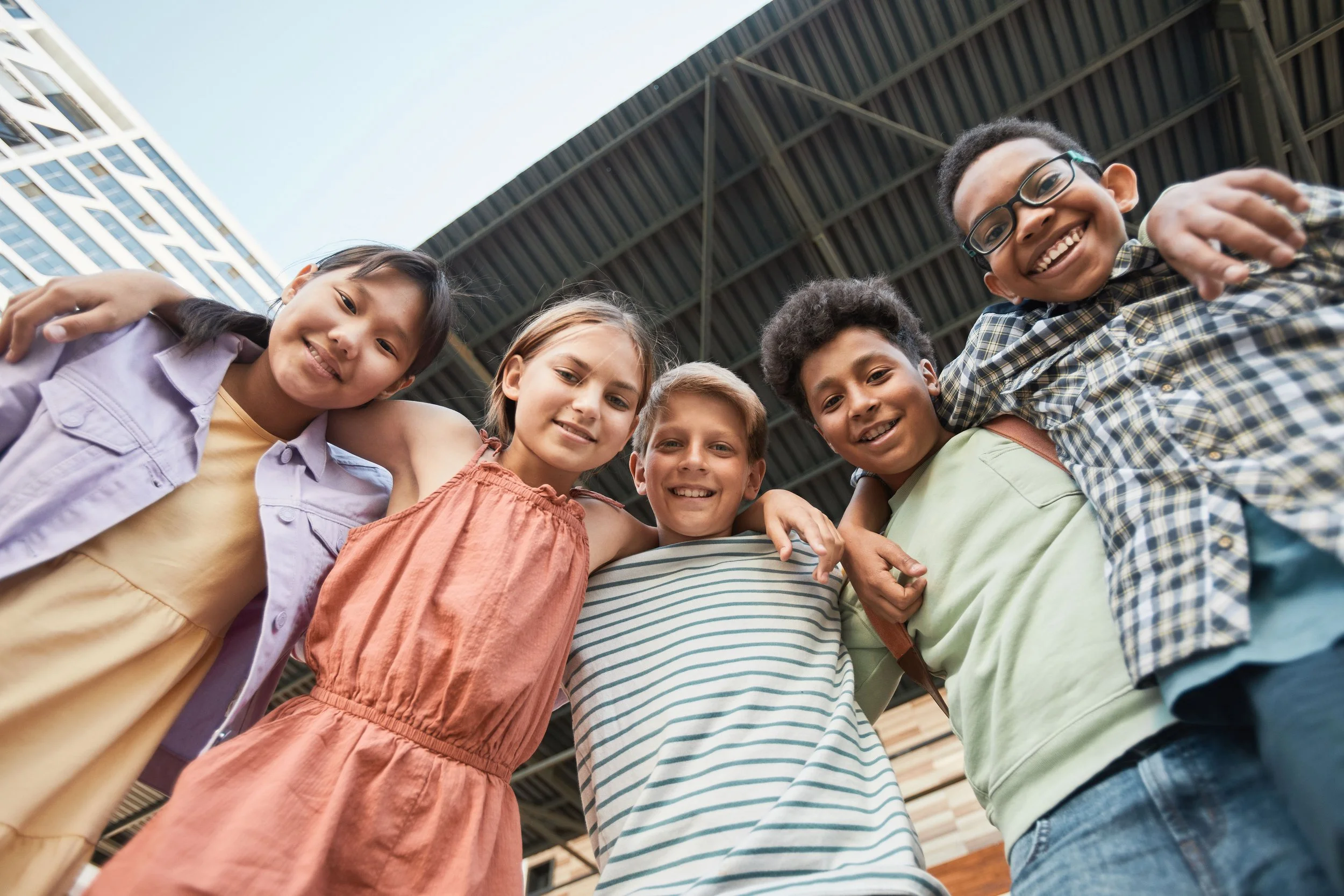 A group of six children smiling and looking down at the camera, standing outside under a large roof structure with a building in the background.