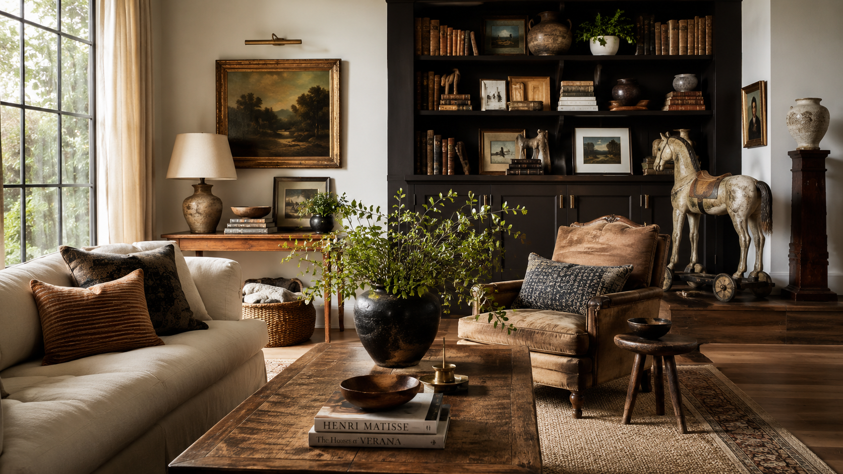 Warm, collected living room with vintage artwork, dark built-in bookshelves, layered books, greenery, and comfortable furnishings.