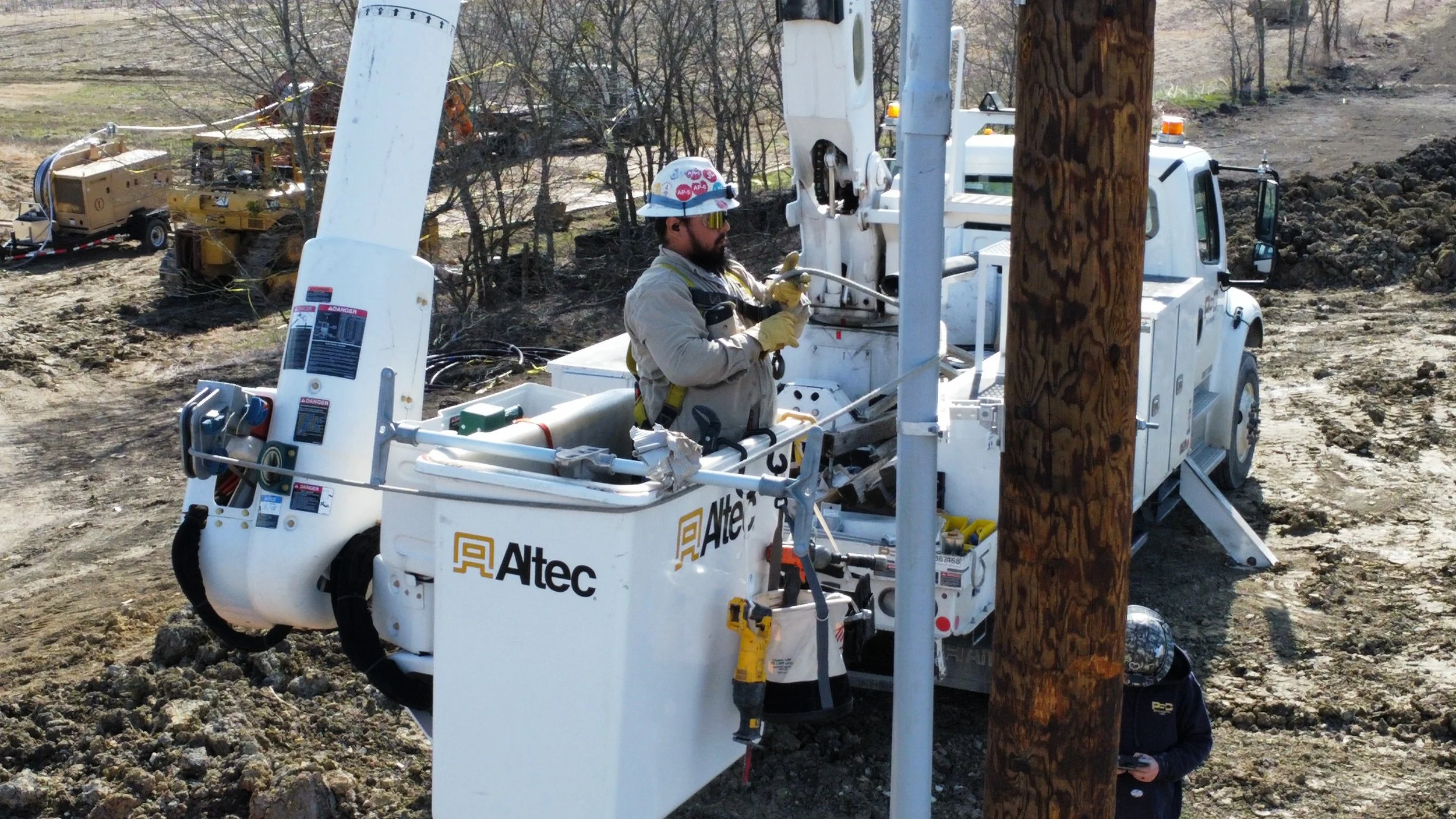 A worker in safety gear operates a bucket truck at a utility pole during daytime, with equipment and machinery in the background on a dirt and rocky terrain.