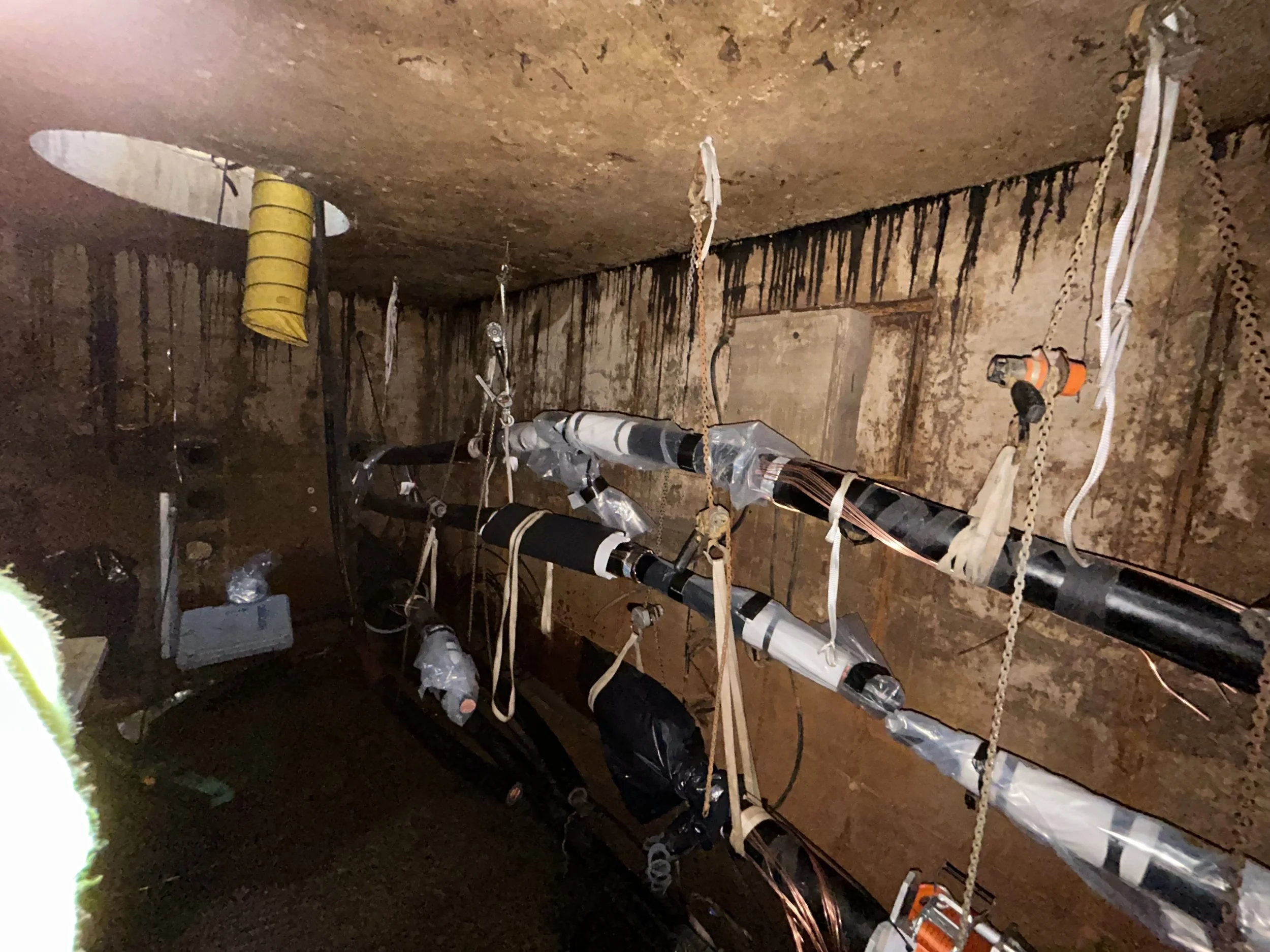 Inside a construction site or underground tunnel showing concrete walls and ceiling with various utility pipes, electrical cables, lights, and safety chains hanging from ceiling.