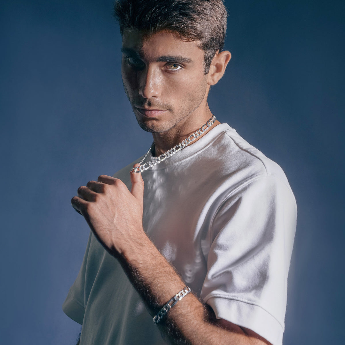 A young man with dark hair and tan skin wearing a white t-shirt, silver jewelry including a chain necklace and bracelet, looking at the camera with a serious expression, against a dark blue background.