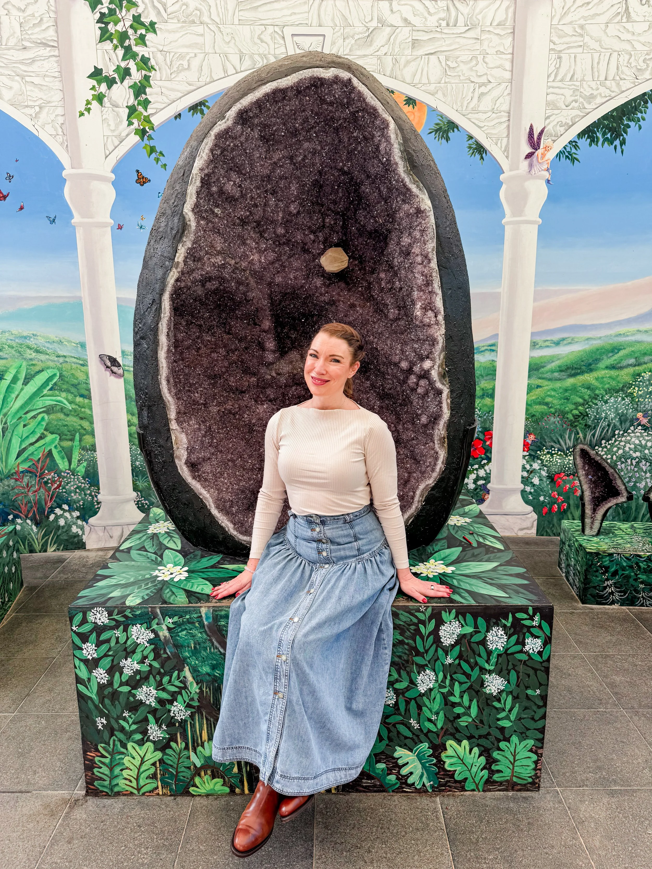 A woman with red hair, wearing a cream-colored sweater and a long blue denim skirt, seated on a decorated bench in front of a large geode stone at a botanical-themed display. The background features a painted mural portraying lush green hills, trees, butterflies, fairies, and arches.