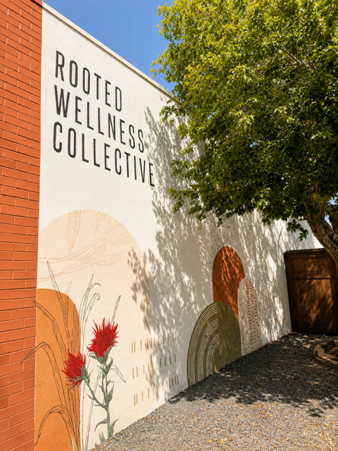 Exterior view of a white building with the words 'Rooted Wellness Collective' on the wall, next to a green tree casting shadows, and colorful artistic shapes at the base of the wall.