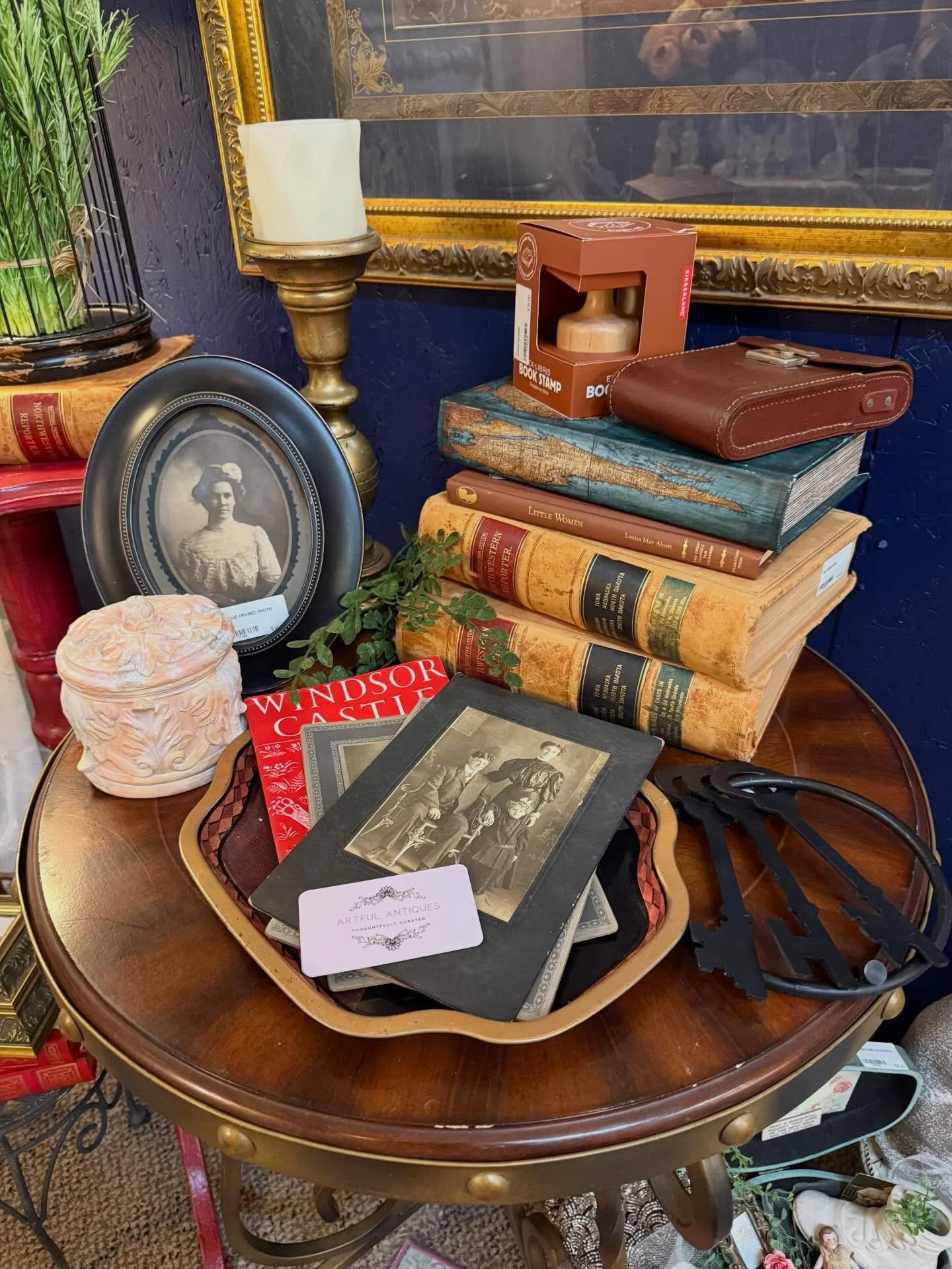 A cluttered wooden table with vintage books, framed photographs, a ceramic container, a tray with jewelry, and various decorative items.