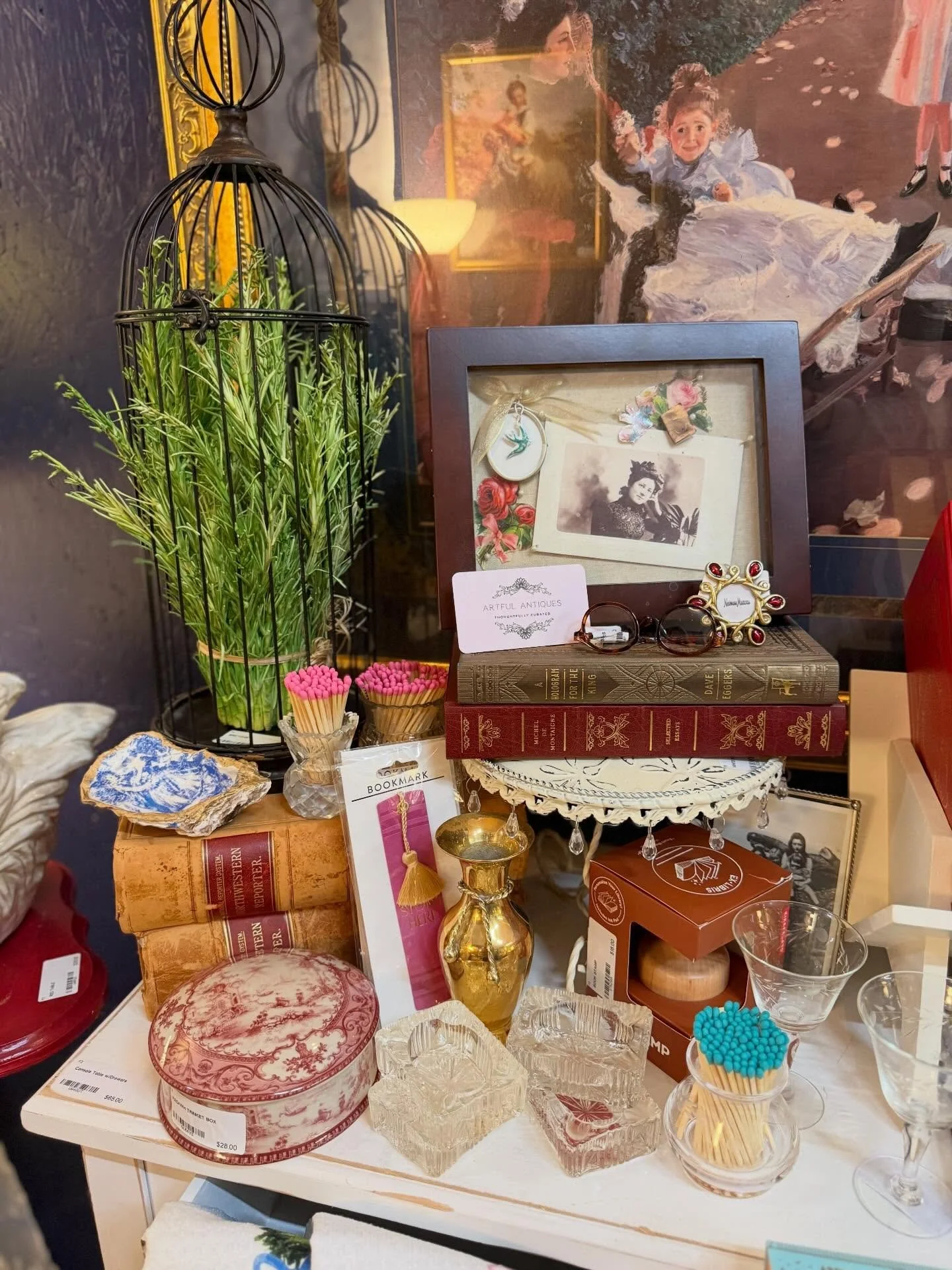 A cluttered vintage decorative display with framed photographs, books, glassware, jewelry, potted plants, and various trinkets on a table.