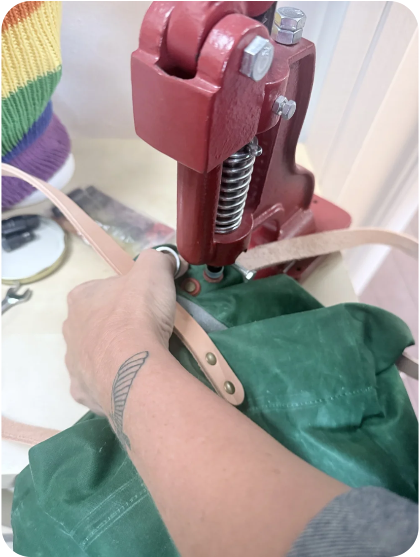 Close-up of a hand adjusting a strap on a green fabric item using a red rivet-pressing machine with rollers and bolts.