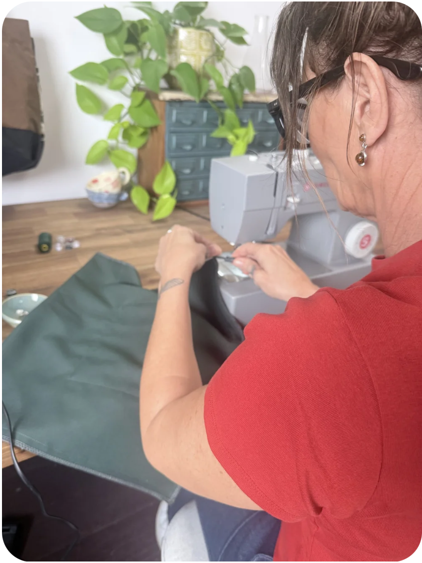 A woman with glasses and earrings sewing fabric at a desk, with tools and plants in the background.
