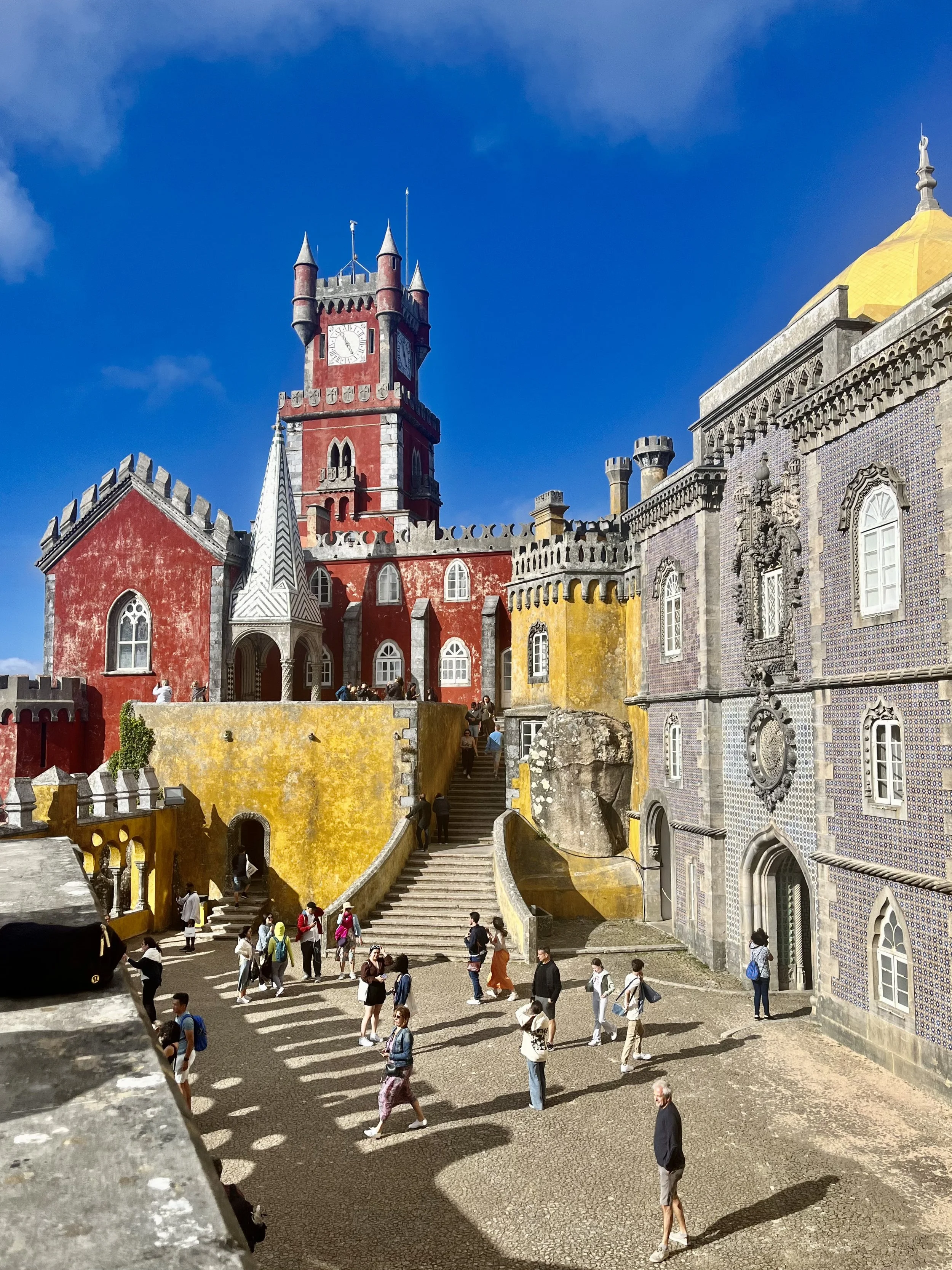 A castle with a red tower, yellow and purple tiled walls, and a clock, filled with tourists walking and taking photos under a blue sky.
