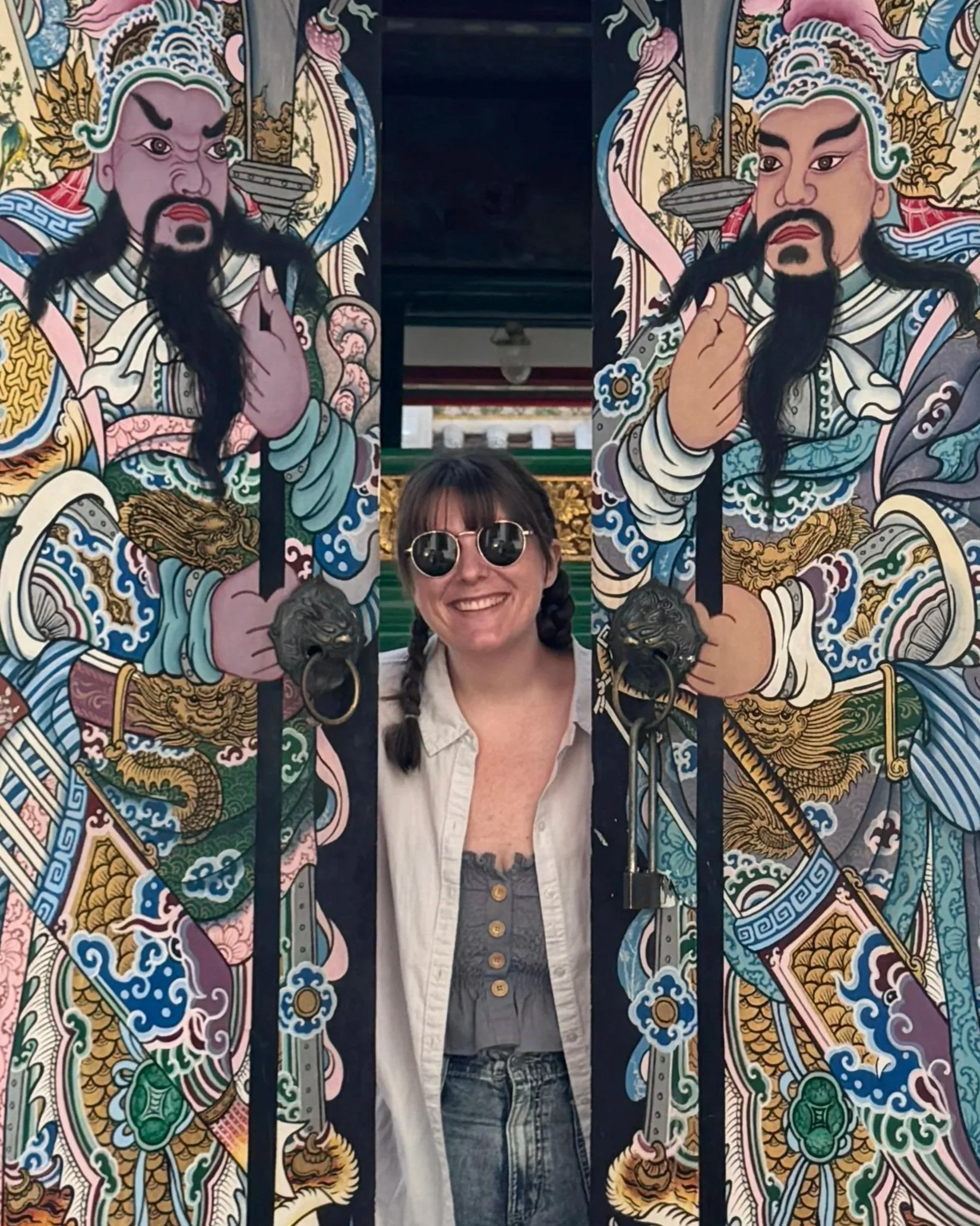 Person with sunglasses smiling behind ornate, colorful Asian-style artwork featuring two fierce warrior figures with long black beards and traditional clothing.