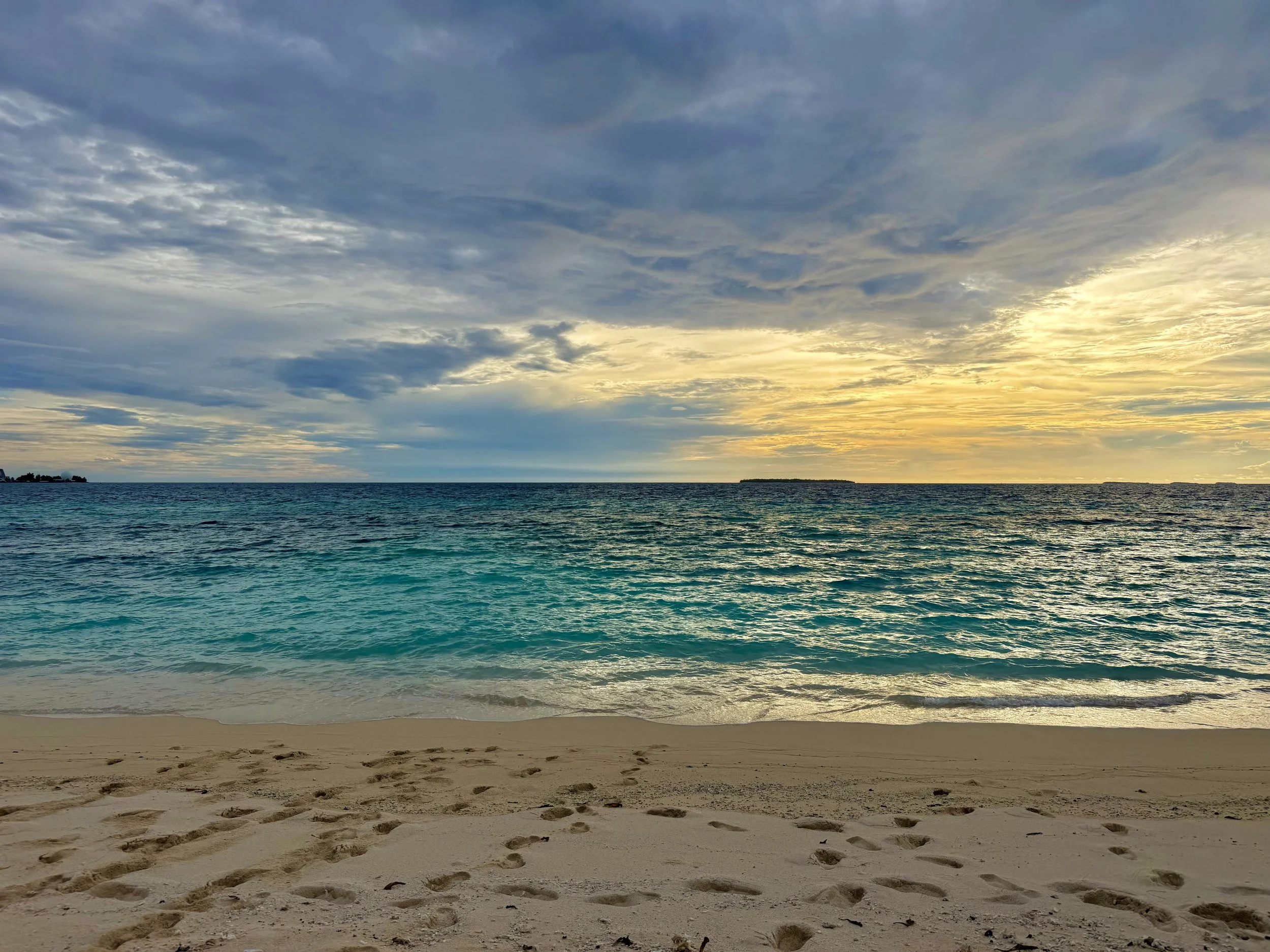 A sandy beach with footprints in the foreground, turquoise water, and a cloudy sky with patches of sunlight breaking through. In the distance, landmasses are visible on the horizon.
