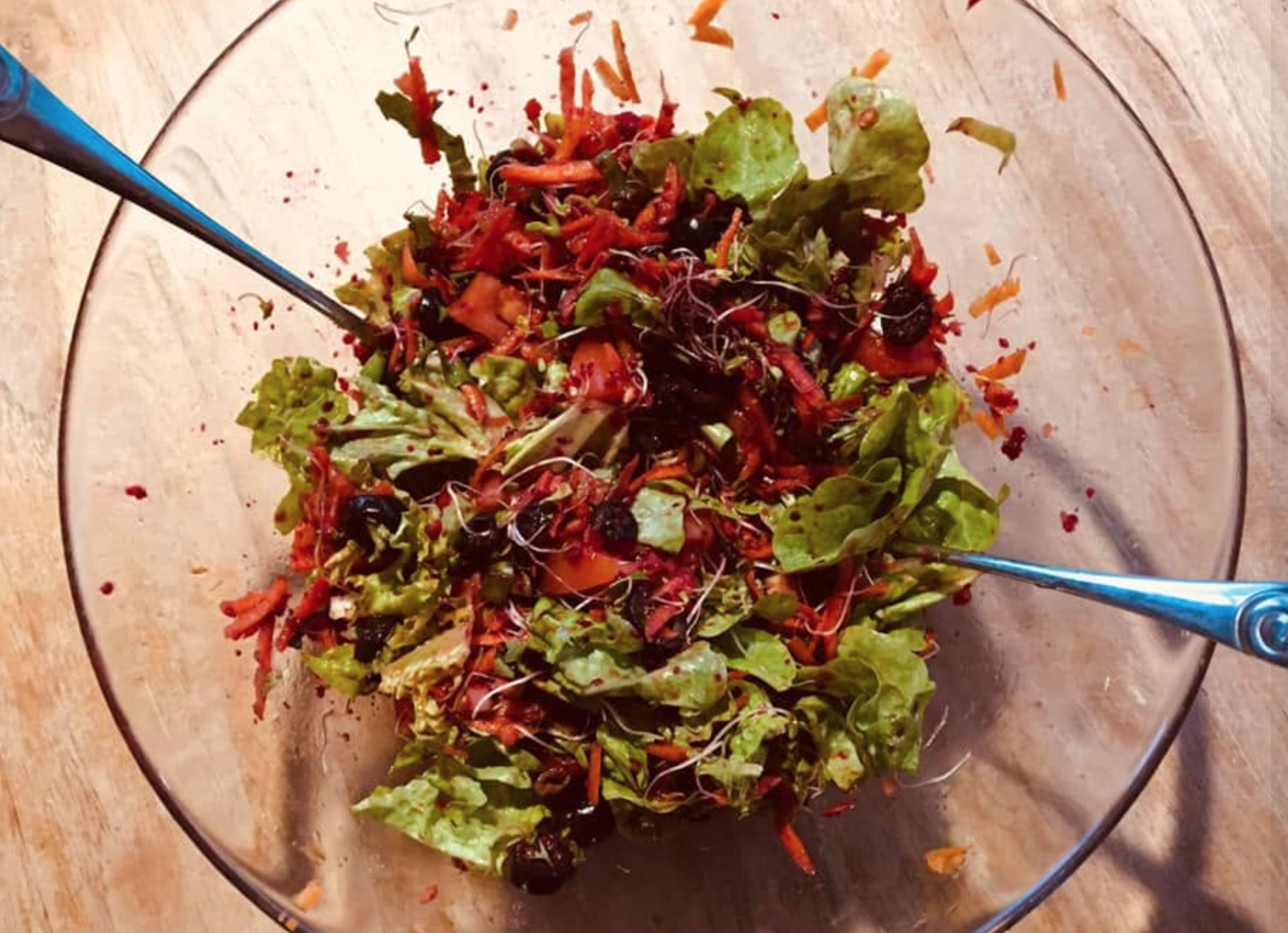 A glass bowl containing a fresh mixed salad with various greens, shredded carrots, and berries, with two metal salad tongs.