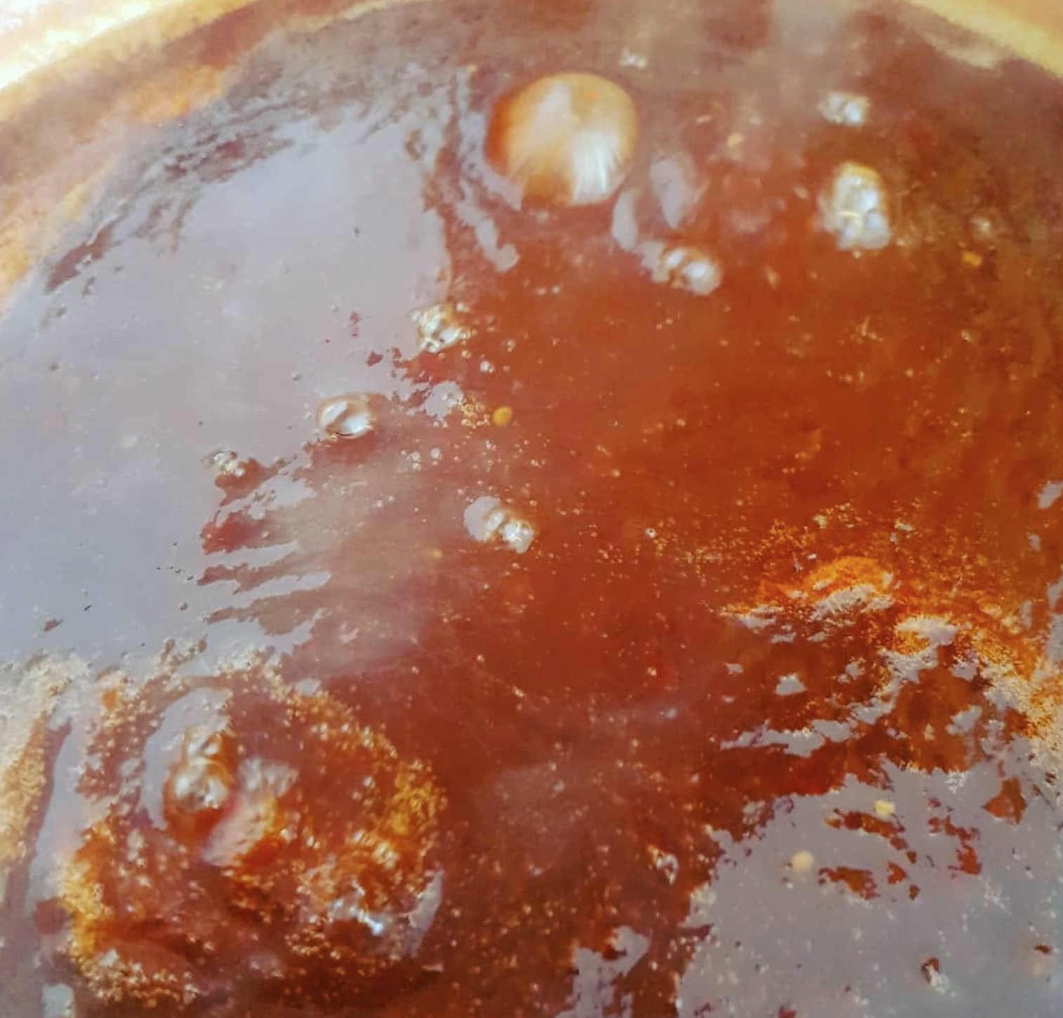 Close-up of a pot with boiling hot, dark red-brown spicy soup or sauce with floating garlic cloves.