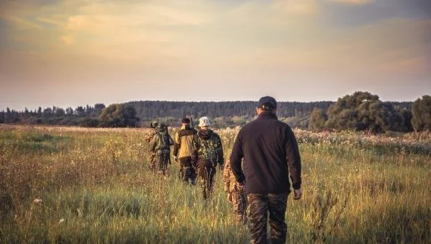 Four people walking through a grassy field at sunset, carrying backpacks.