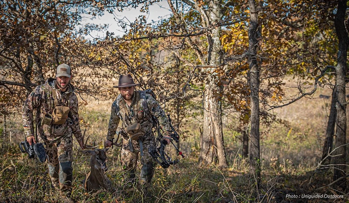 Two hunters in camouflage clothing pose in a wooded area with a deer they have hunted, holding its antlers. They are carrying hunting equipment, and one is holding a camera. The scene is set in fall with trees in the background.