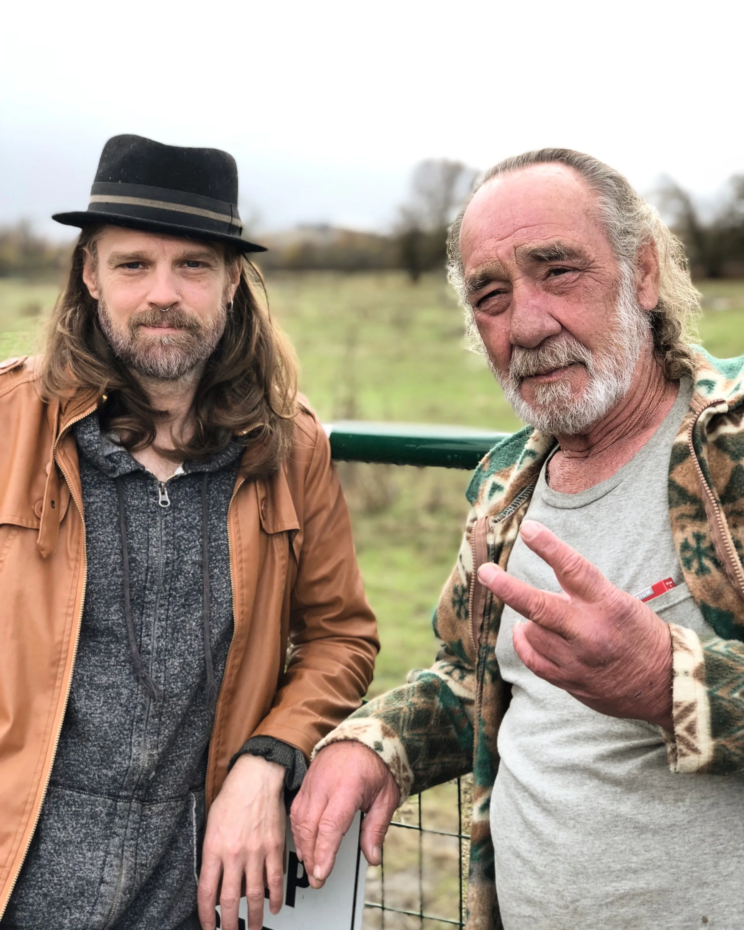 Two men standing outdoors, one with long hair and a beard wearing a fedora hat and brown jacket, the other with gray hair and beard making a peace sign, both near a fence on an overcast day.