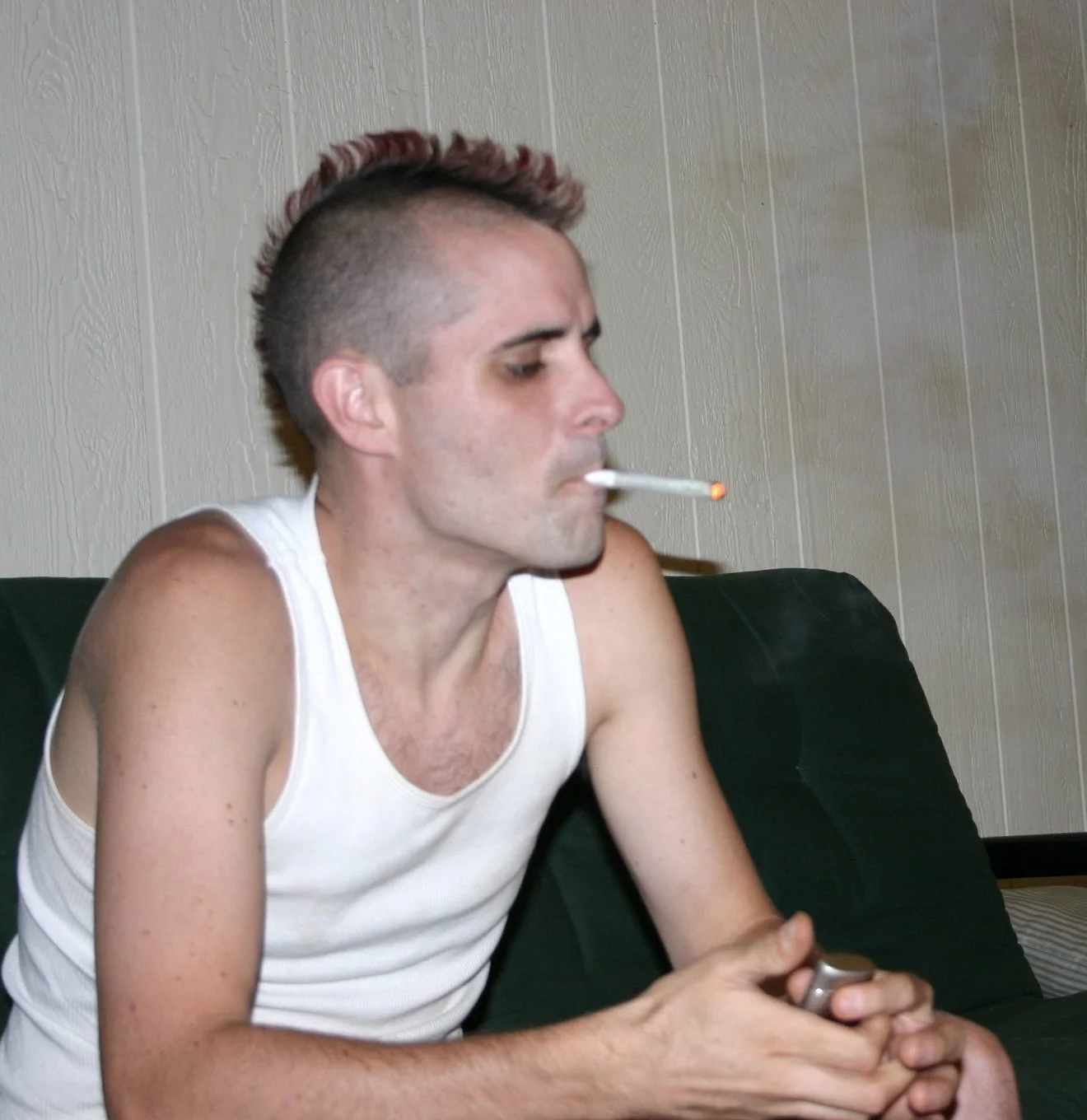 Young man with a mohawk hairstyle, wearing a white tank top, smoking a cigarette, sitting on a dark green couch, holding a phone, in a room with light-colored wood-paneled walls.