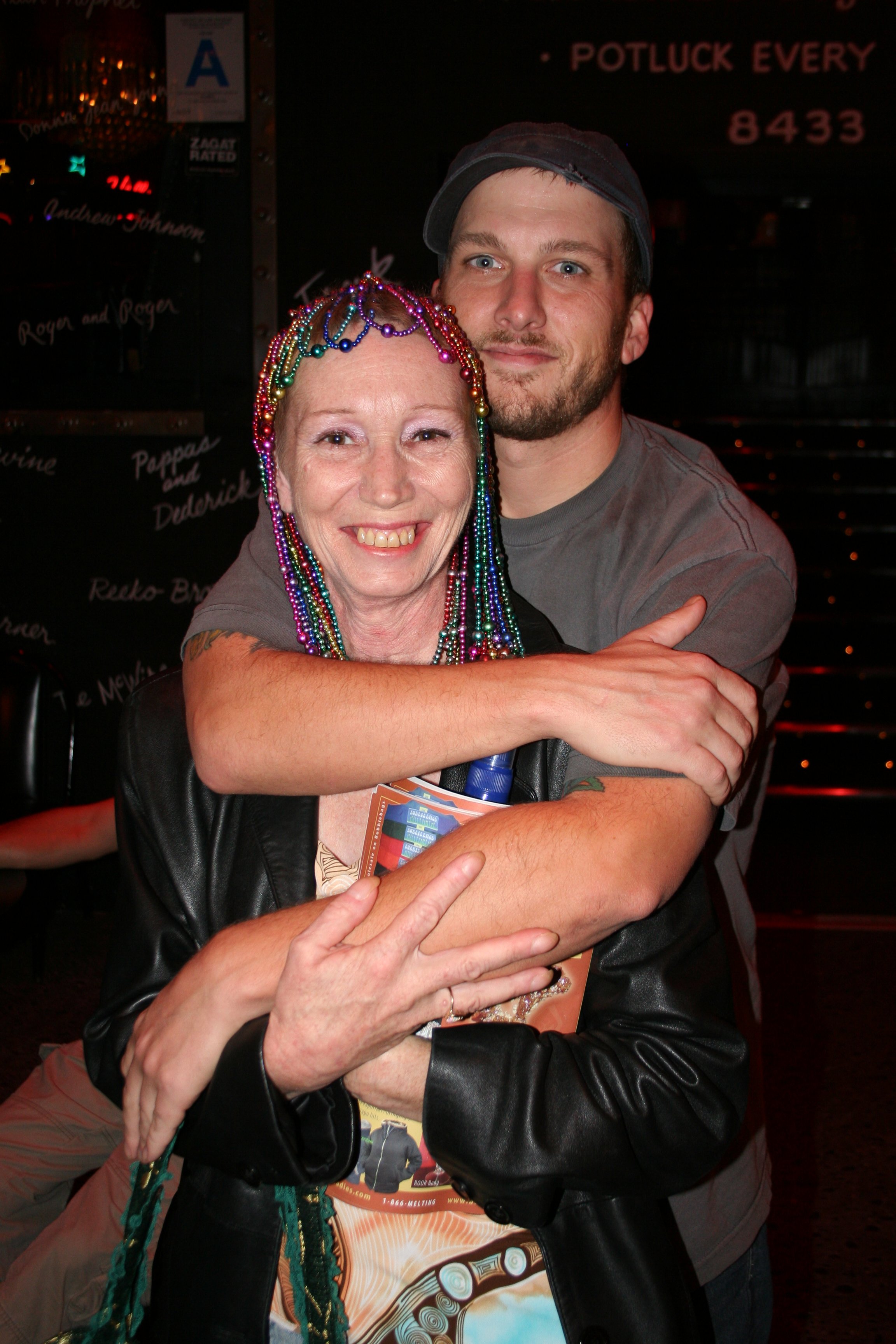 A woman with colorful mardi gras beads and a man in a gray t-shirt and baseball cap hugging in a bar or pub.