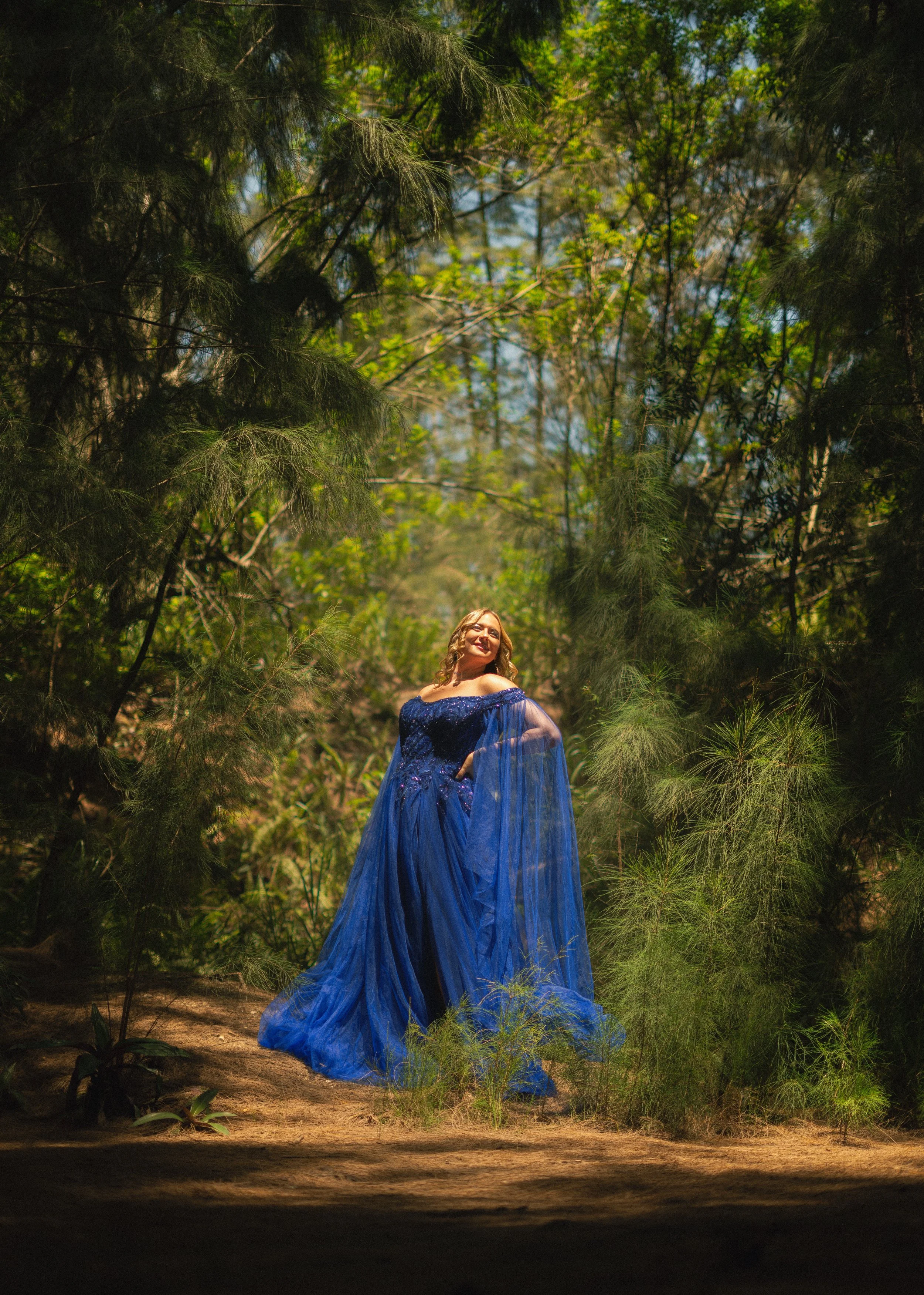 Woman in a flowing blue gown standing outdoors amidst tall trees and green foliage.