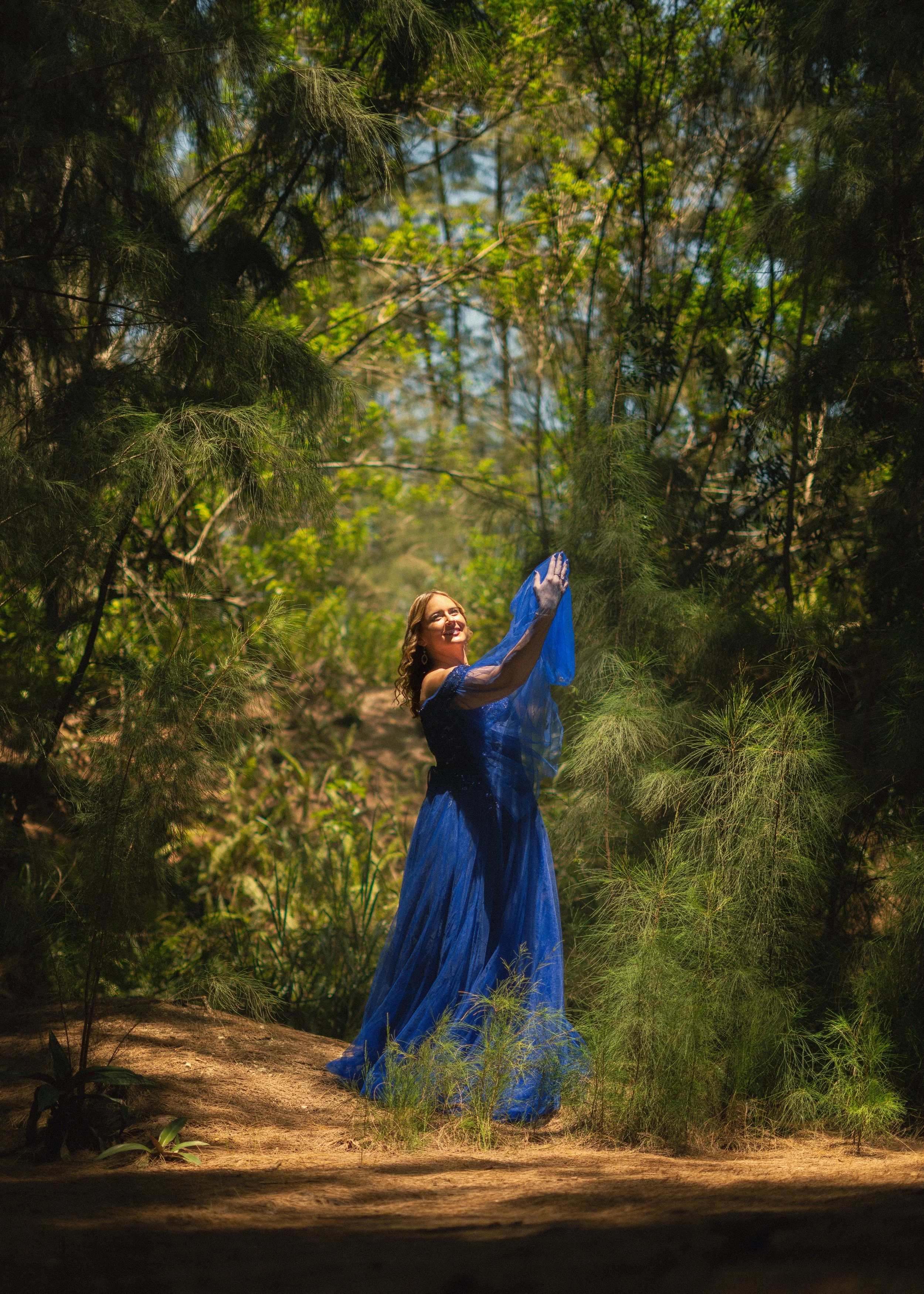 A woman in a long blue dress standing among green and brown trees, holding a blue sheer cloth, looking upward, in a forest setting with sunlight filtering through the trees.