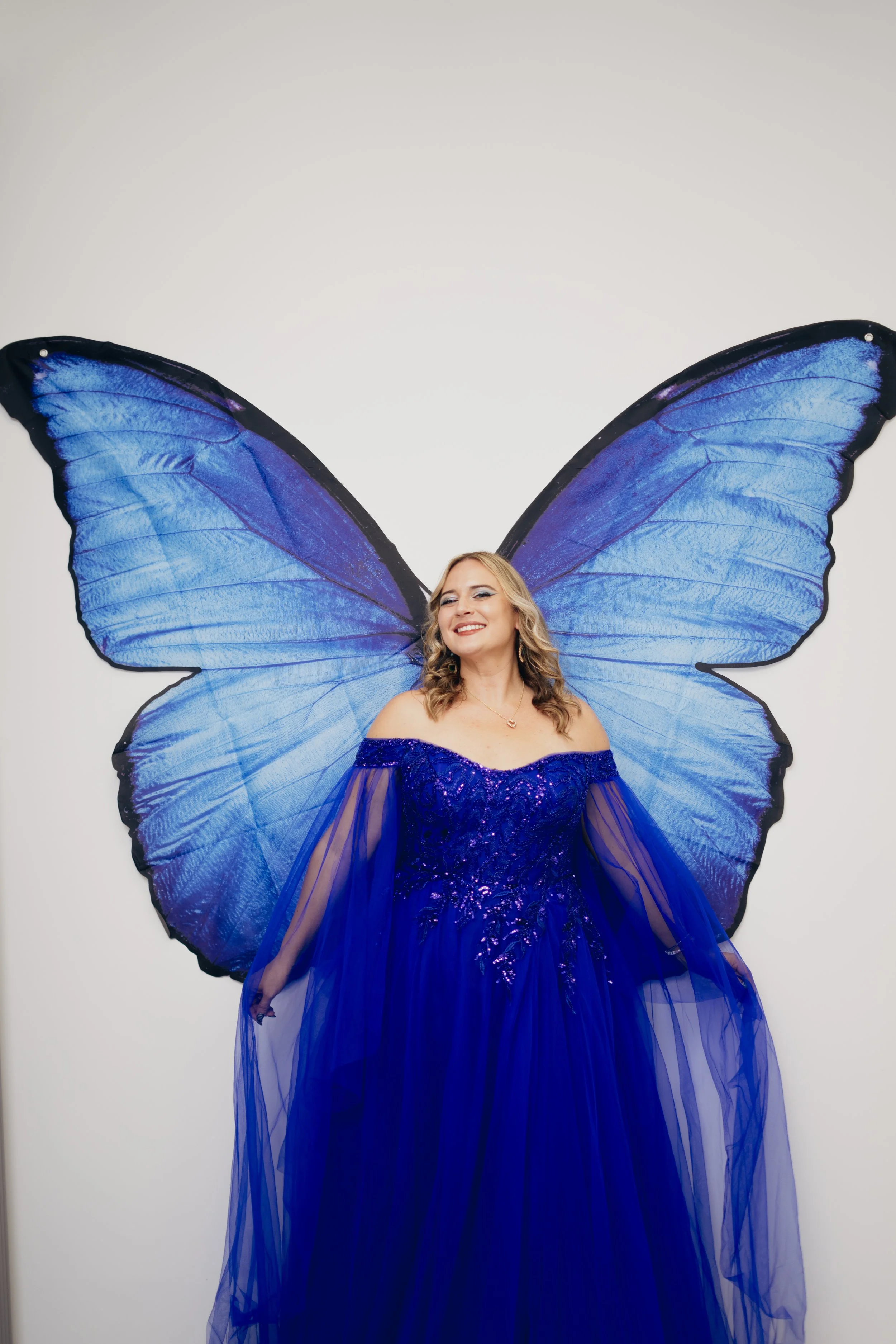 A woman dressed in a royal blue gown with sheer fabric and sequins, smiling and posing in front of a large butterfly wing backdrop.