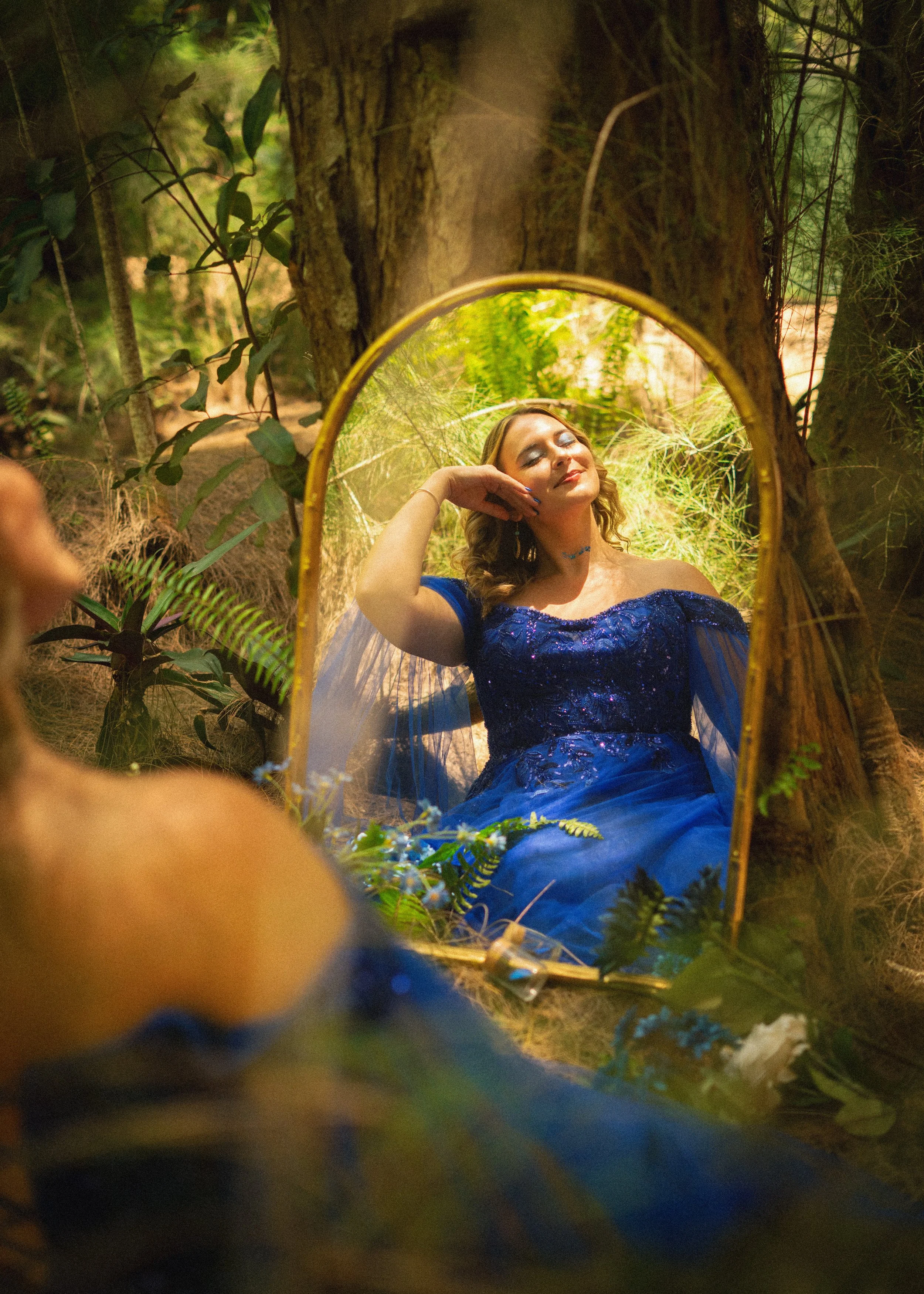 A woman in a blue dress with makeup and jewelry, sitting with her eyes closed and smiling, looking into a mirror in a forest setting surrounded by greenery, trees, and branches.