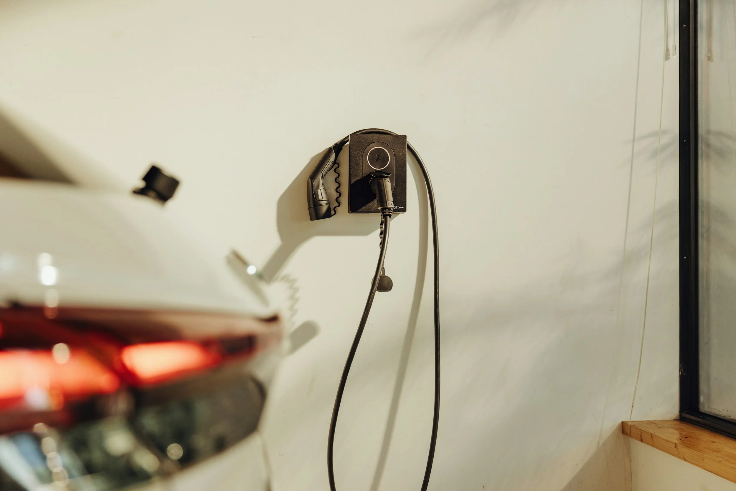A black wall-mounted charging station with a cord and plug, situated on a plain, light-colored wall. Part of a blurred red object appears in the foreground.