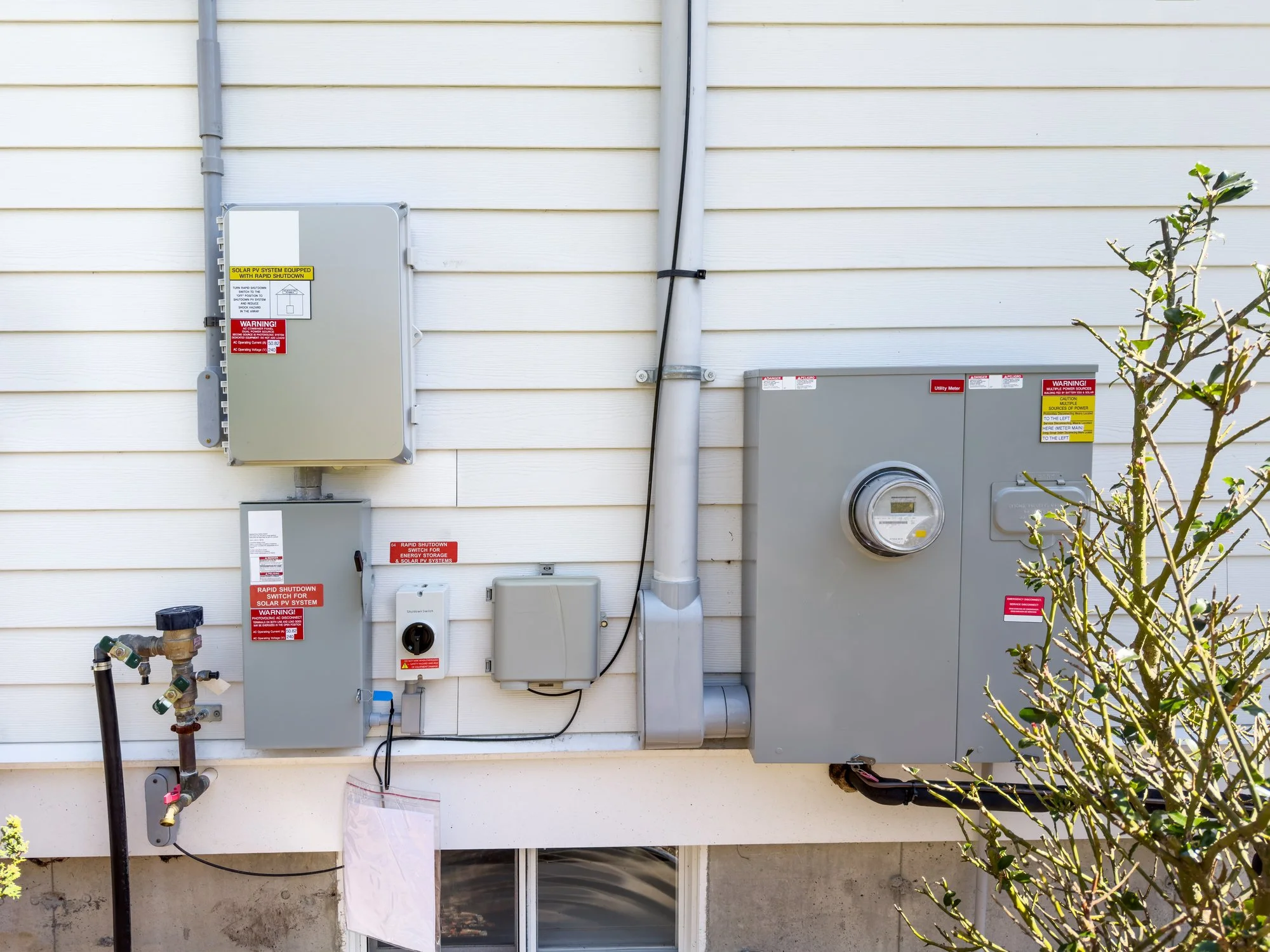Exterior wall with electrical and solar power equipment, including a metal meter box, electrical panels, warning labels, and conduit pipes, with a small bush partially visible on the right.