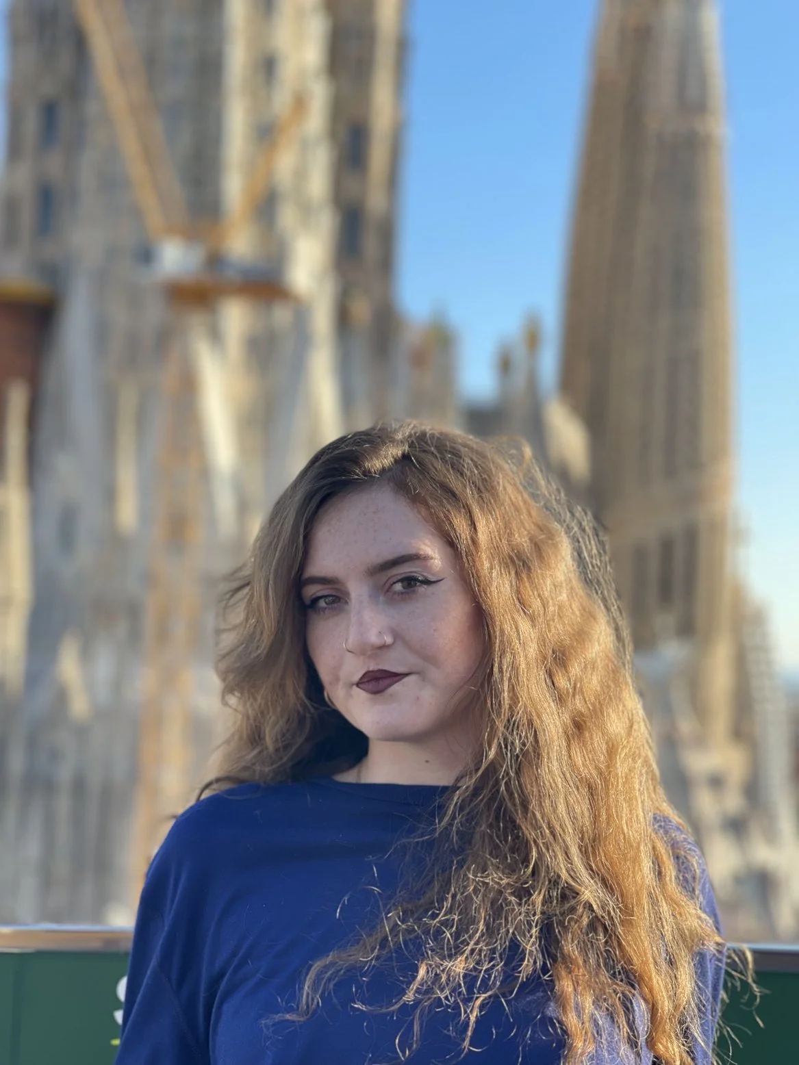 A young woman with long, wavy red hair and dark lipstick posing in front of the Sagrada Familia basilica in Barcelona, Spain, during daylight.