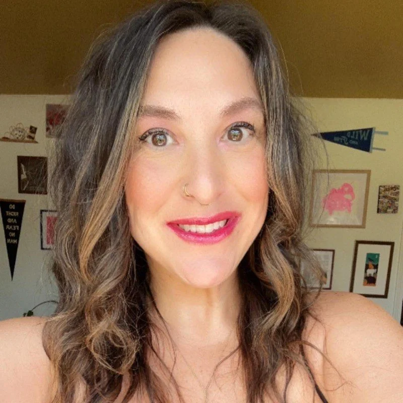 Close-up of smiling woman with wavy brown hair, wearing a gold nose ring and pink lipstick, in a room with framed pictures and decorations on the wall.