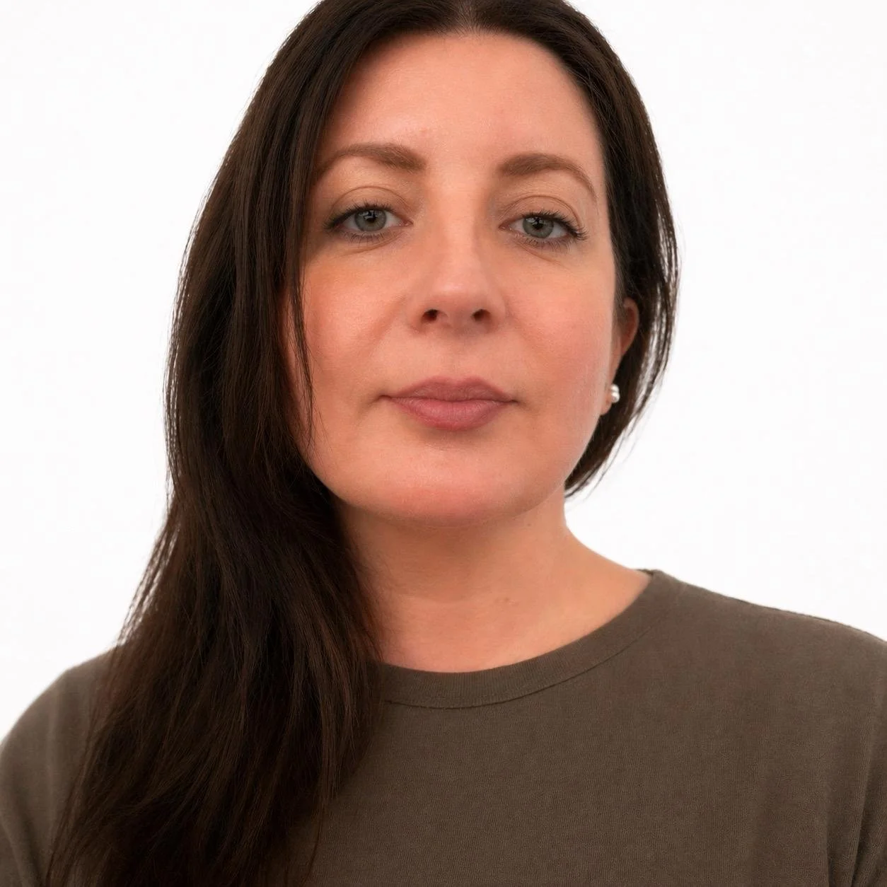 A woman with long dark hair, light skin, and blue eyes, wearing a brown top and pearl earrings, posed in front of a white background.