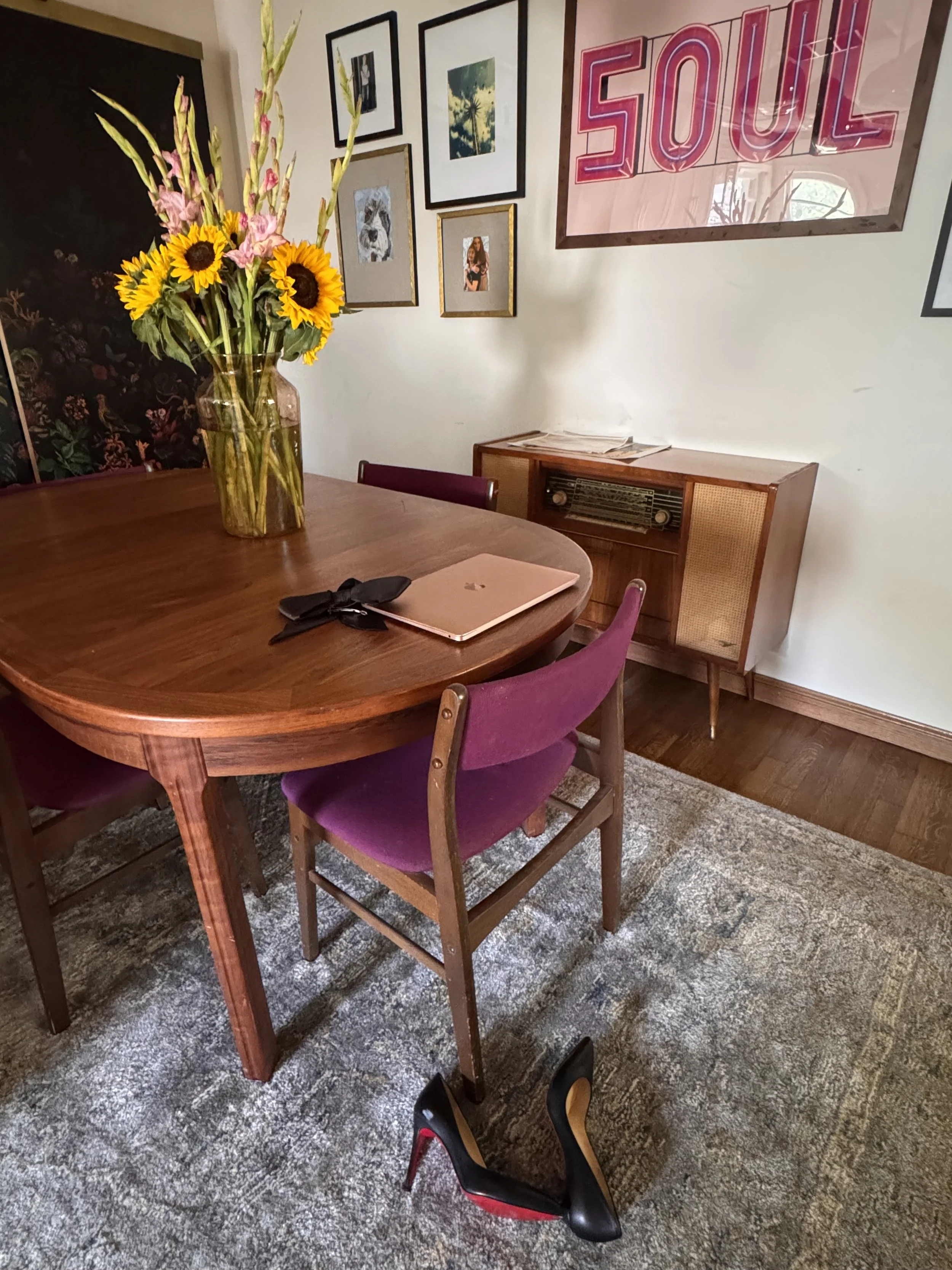 A dining room with a wooden table with a leather-bound notebook, black glasses, and purple chairs surrounding it. A large glass vase with sunflowers and pink gladiolus on the table. In the background, a vintage radio cabinet, framed art on the wall, and a large framed sign reading 'SOUL' are visible. The room features a patterned area rug and hardwood flooring.