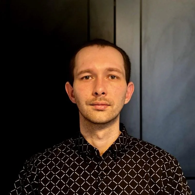 A young man with short brown hair and light skin, wearing a black shirt with a white geometric pattern, standing indoors against a dark wall.