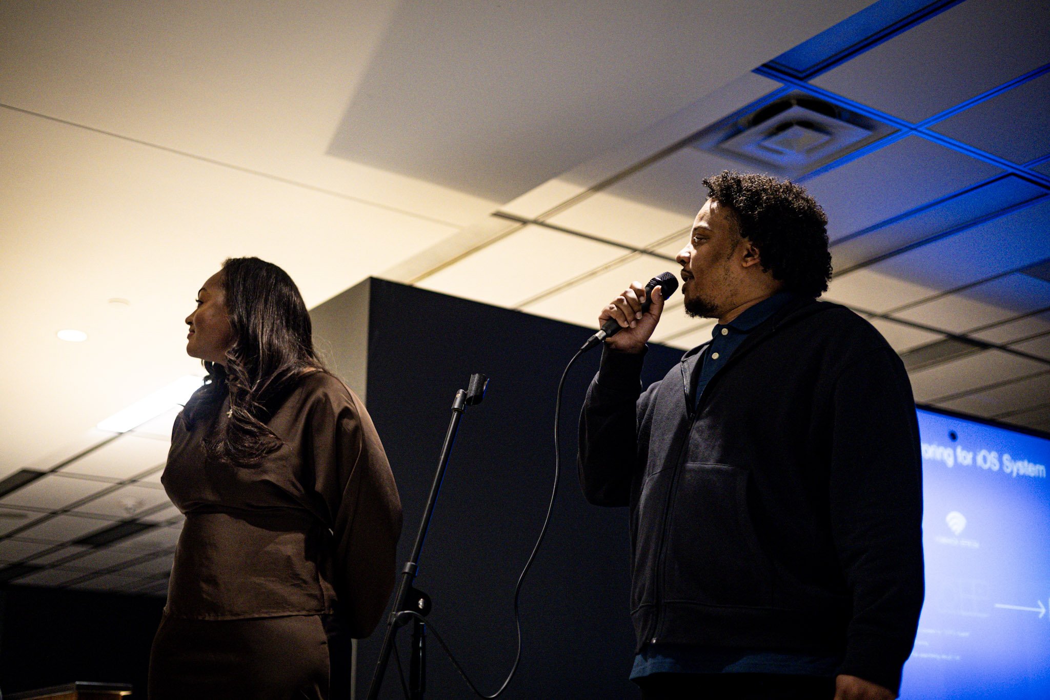A man holding a microphone speaking on stage next to a woman standing with hands behind her back, with a presentation screen displaying partially visible text in the background.