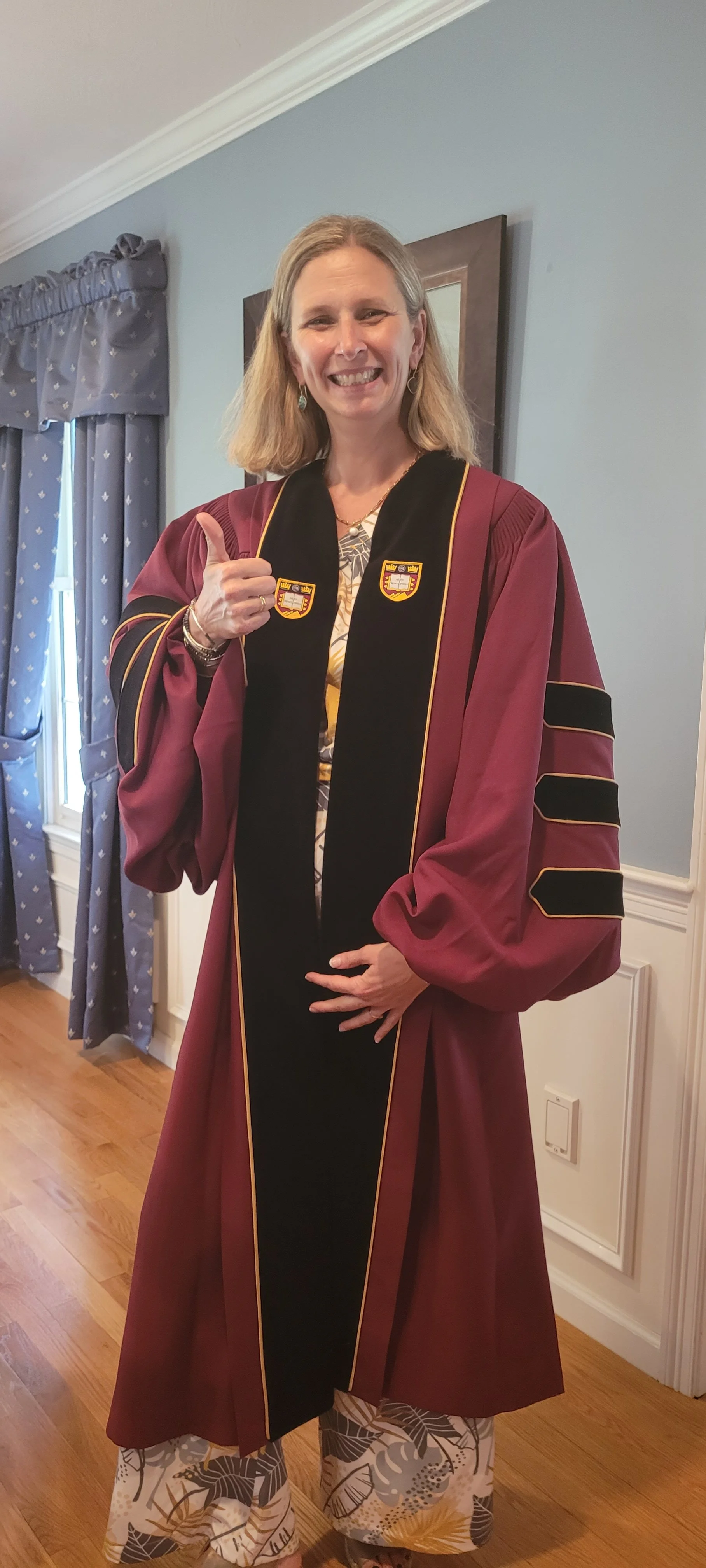 Woman in academic gown giving a thumbs-up in a room with blue walls and wooden flooring.
