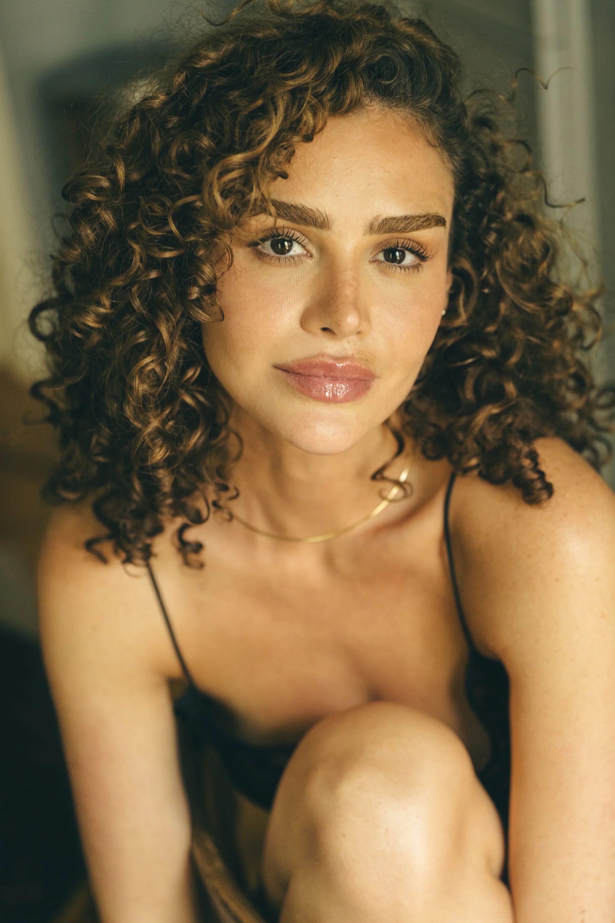 A young woman with curly brown hair, light skin, and wearing a black spaghetti strap top, looks into the camera with a slight smile.