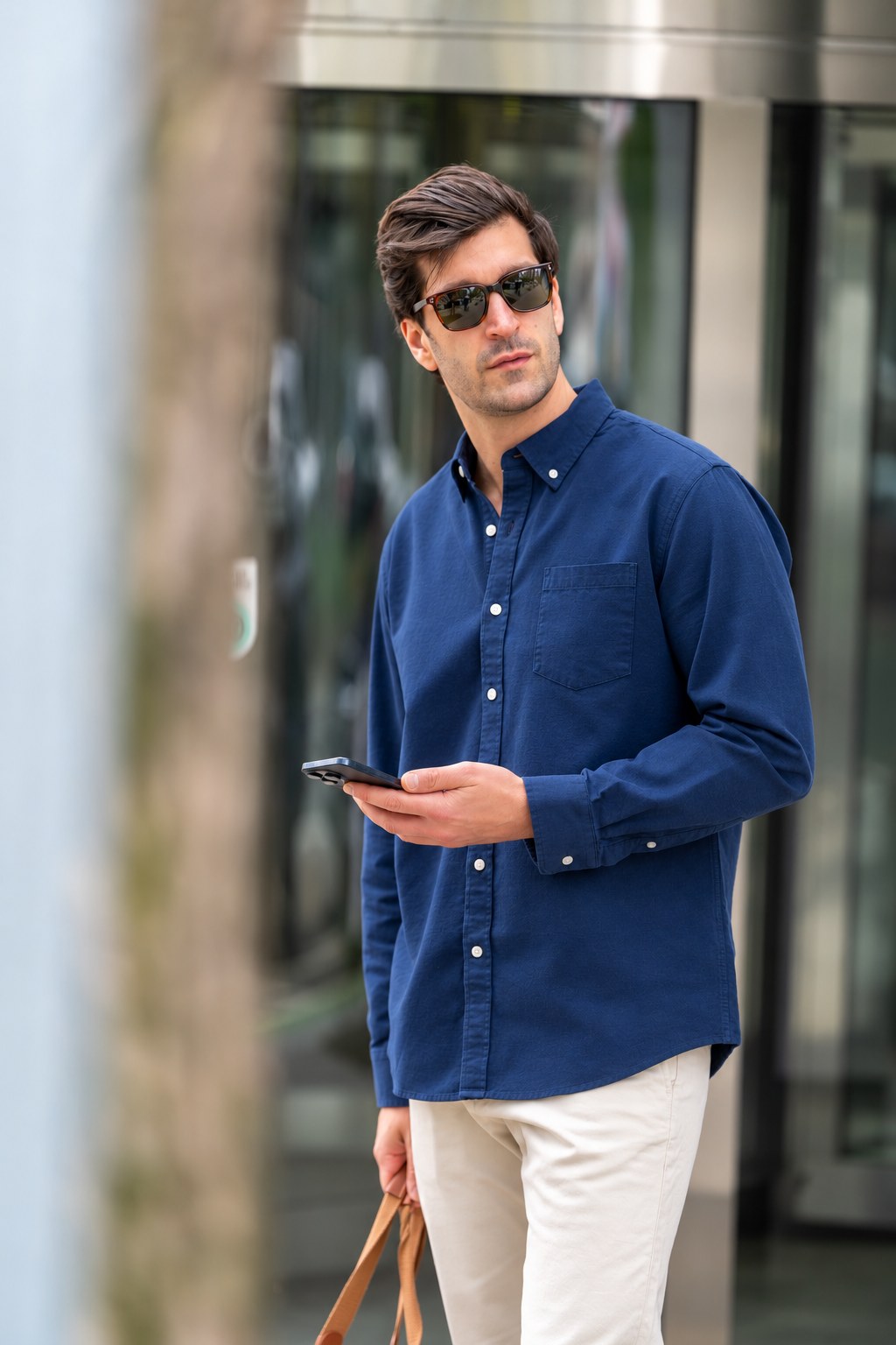 Man wearing sunglasses, a navy blue button-up shirt, and white pants holding a smartphone and a brown bag, outdoors in front of a building.
