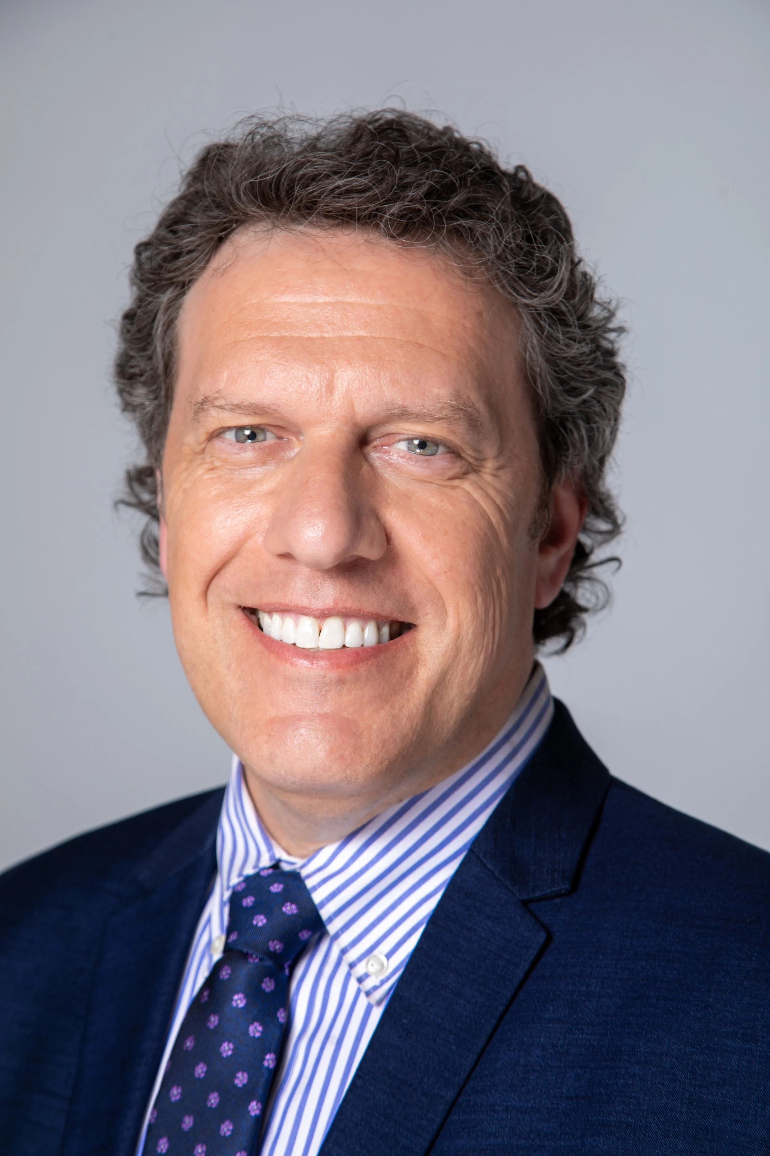 Close-up of a smiling man wearing a dark suit, blue and white striped shirt, and a dark tie with purple floral pattern. Gray background.