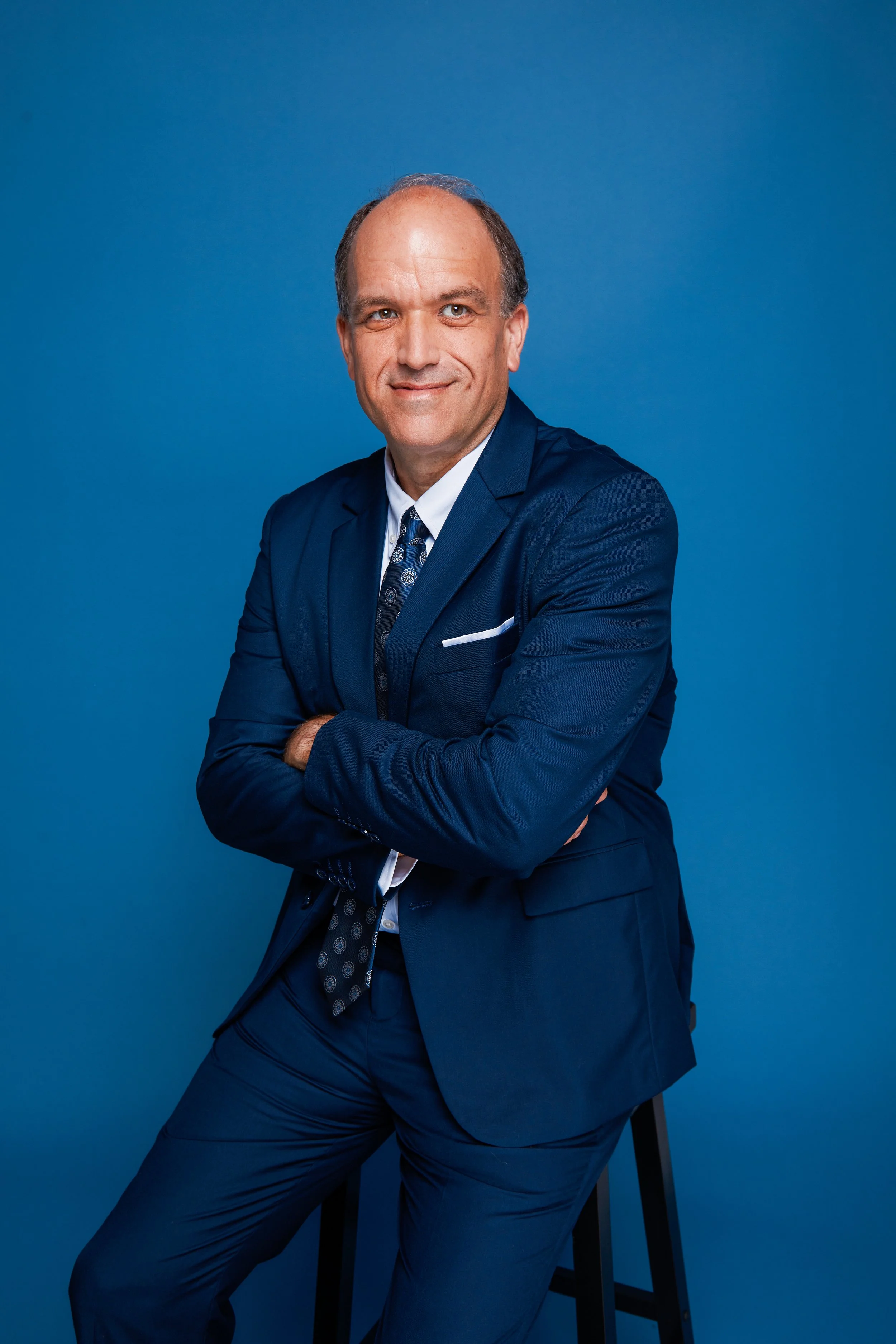 A man in a navy blue suit with a patterned tie and white shirt, smiling with arms crossed, sitting on a black stool against a solid blue background.