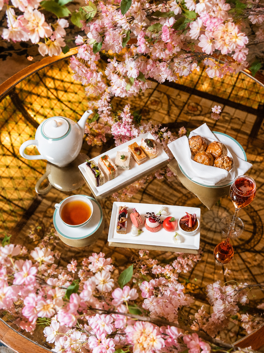 An elegant tea party setup with assorted desserts, a teapot, tea cup, and drinks on a glass table surrounded by pink cherry blossoms.