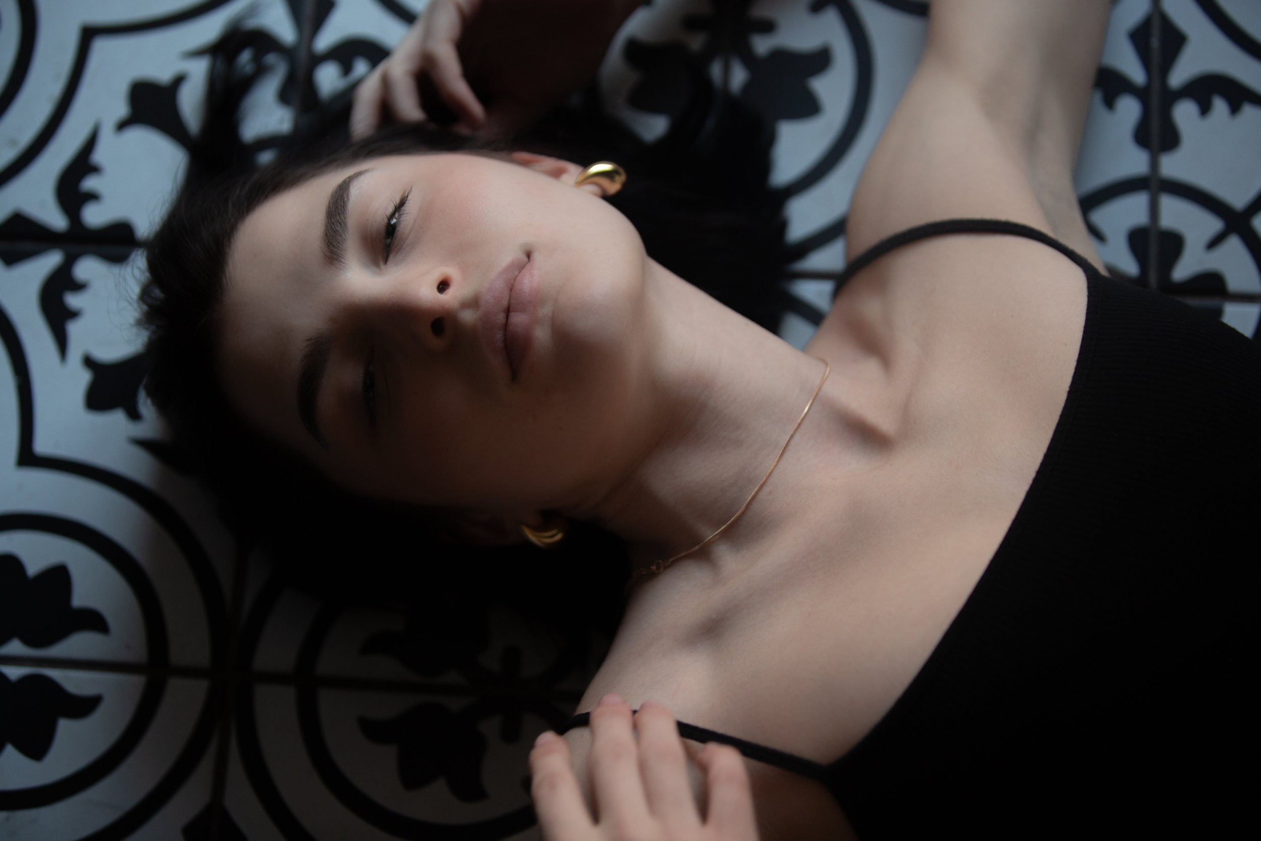 A woman with dark hair lies on a decorative black and white patterned surface, eyes closed, wearing gold hoop earrings and a black top with diagonal strap details.
