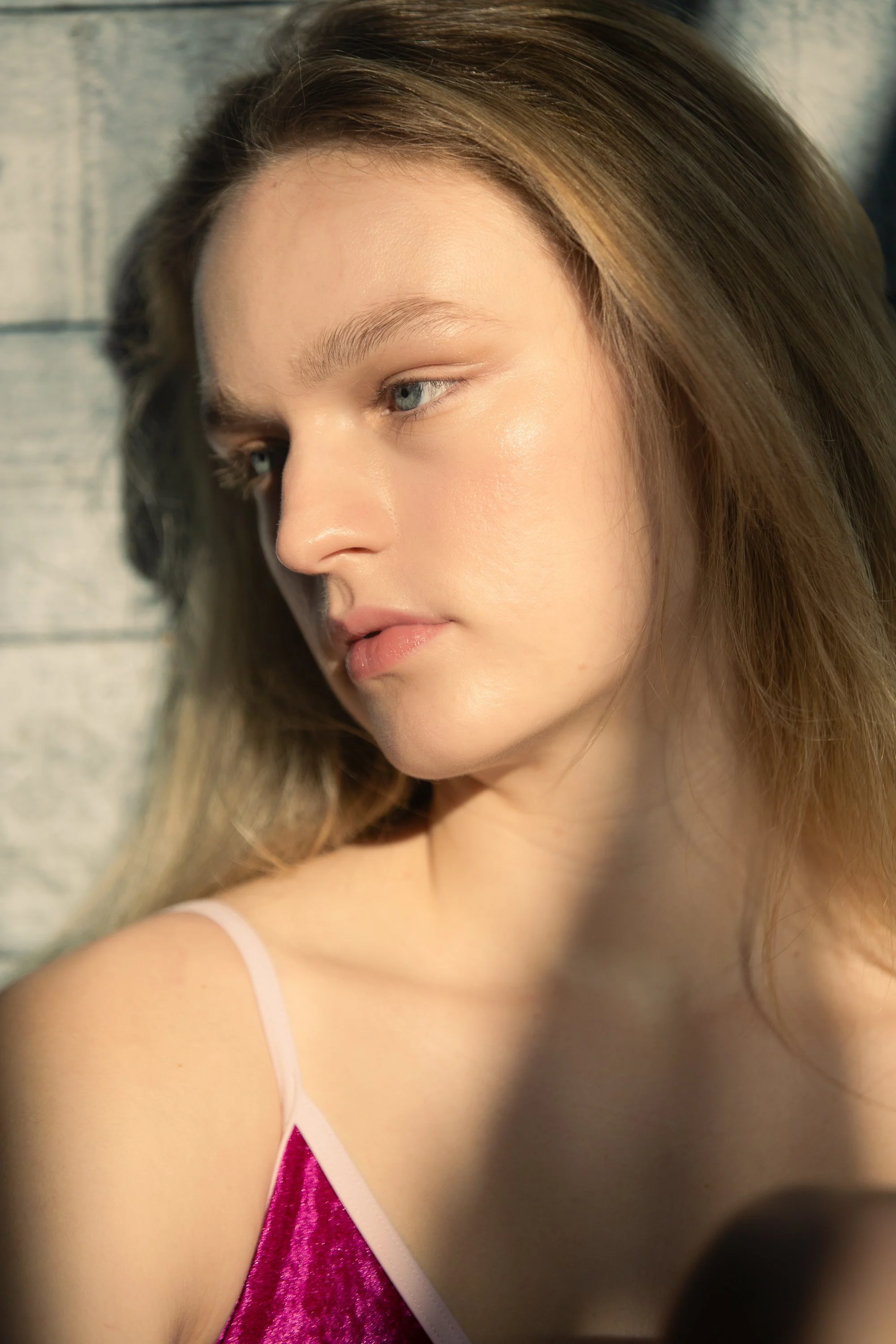Close-up of a young woman with light skin, light brown hair, and blue eyes, wearing a pink satin camisole with white straps, looking down and to the side, with a soft focus background.