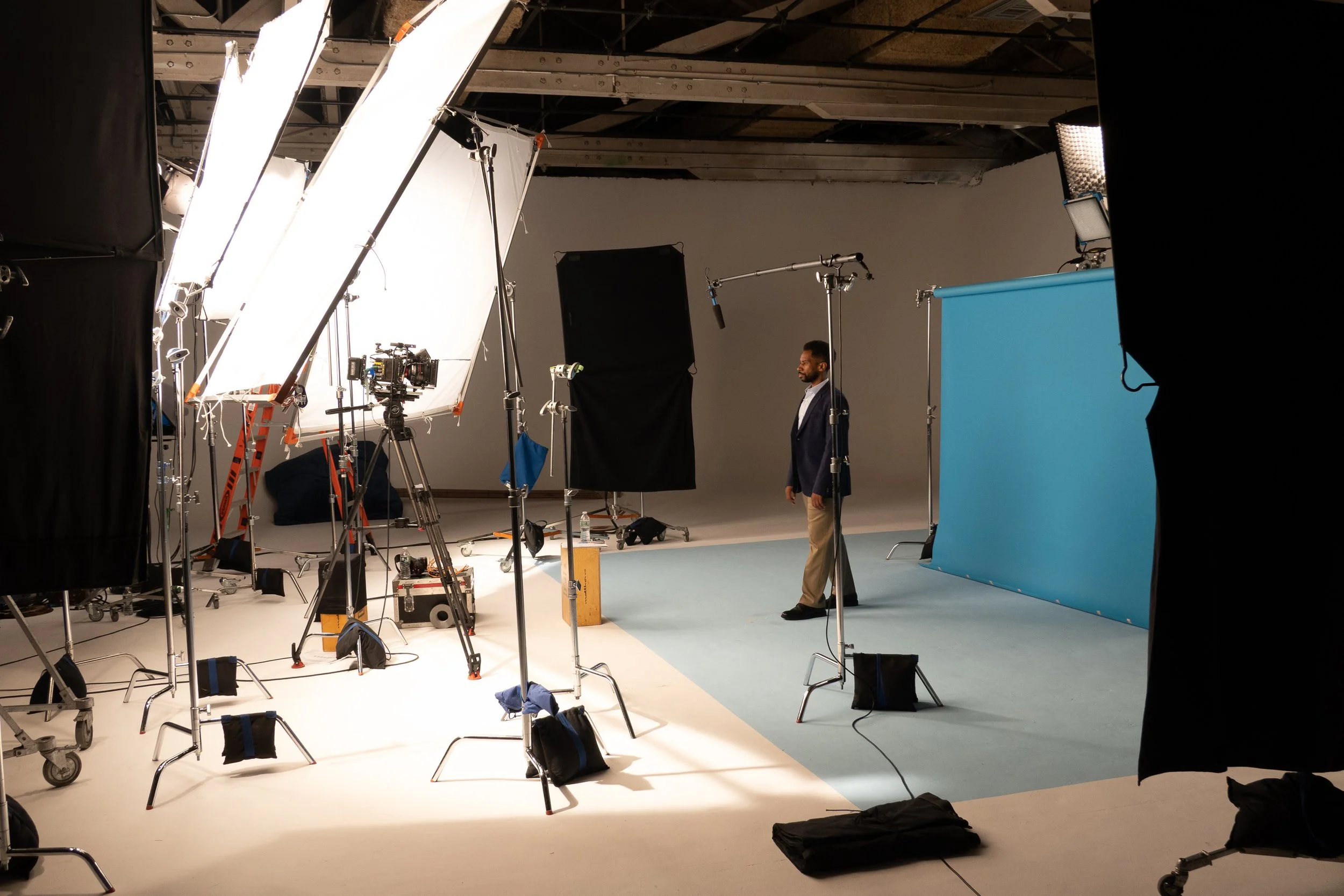 A professional filming studio with lighting equipment, cameras, and a man in a suit standing near a blue screen backdrop.