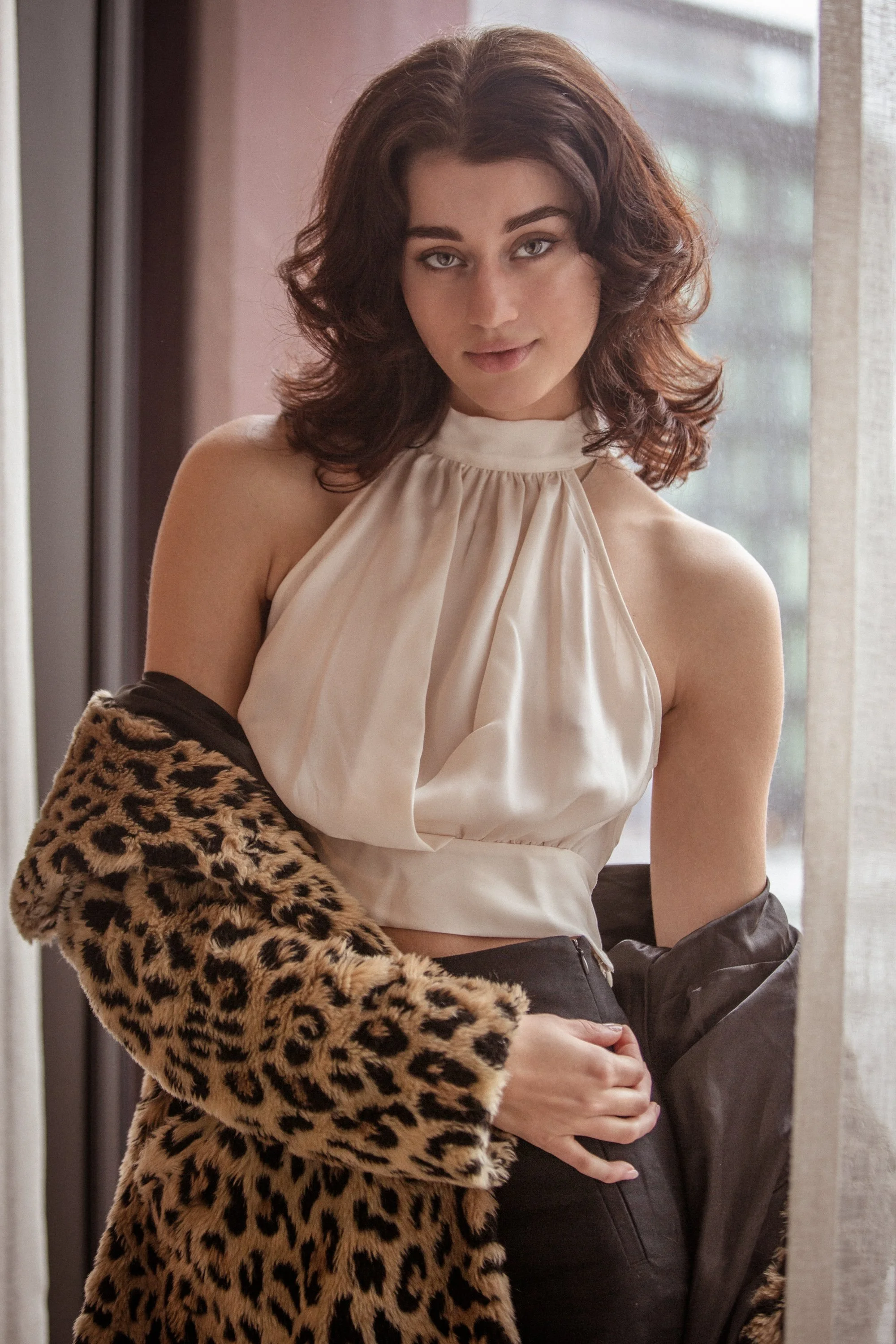 A woman with shoulder-length brown hair, wearing a sleeveless cream-colored top, a faux fur leopard print coat, and black pants, standing near a window with a light-colored curtain.