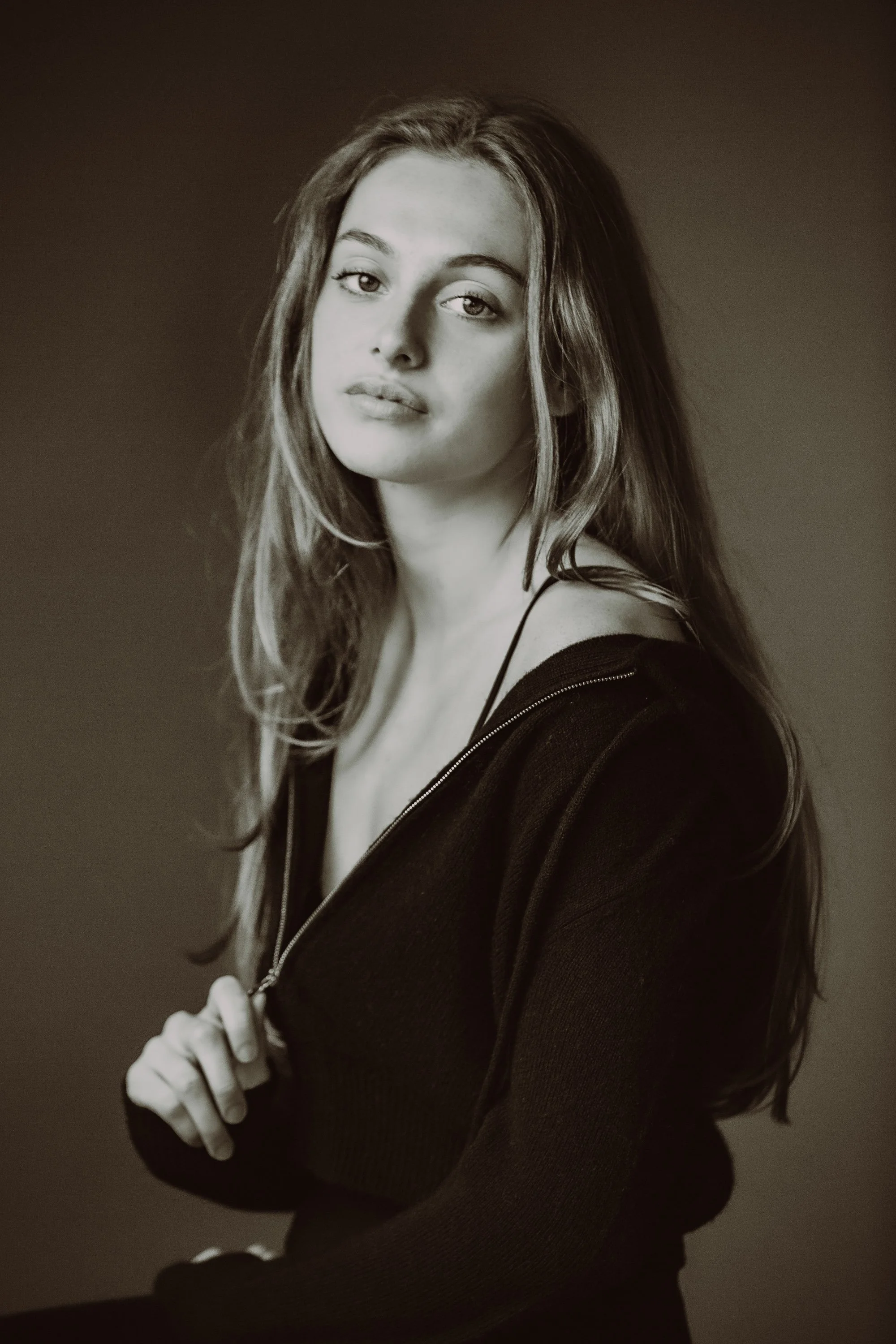 Black and white portrait of a young woman with long wavy hair, wearing a zip-up jacket and a tank top, posing with her body turned slightly to the side and her face looking at the camera, against a plain background.