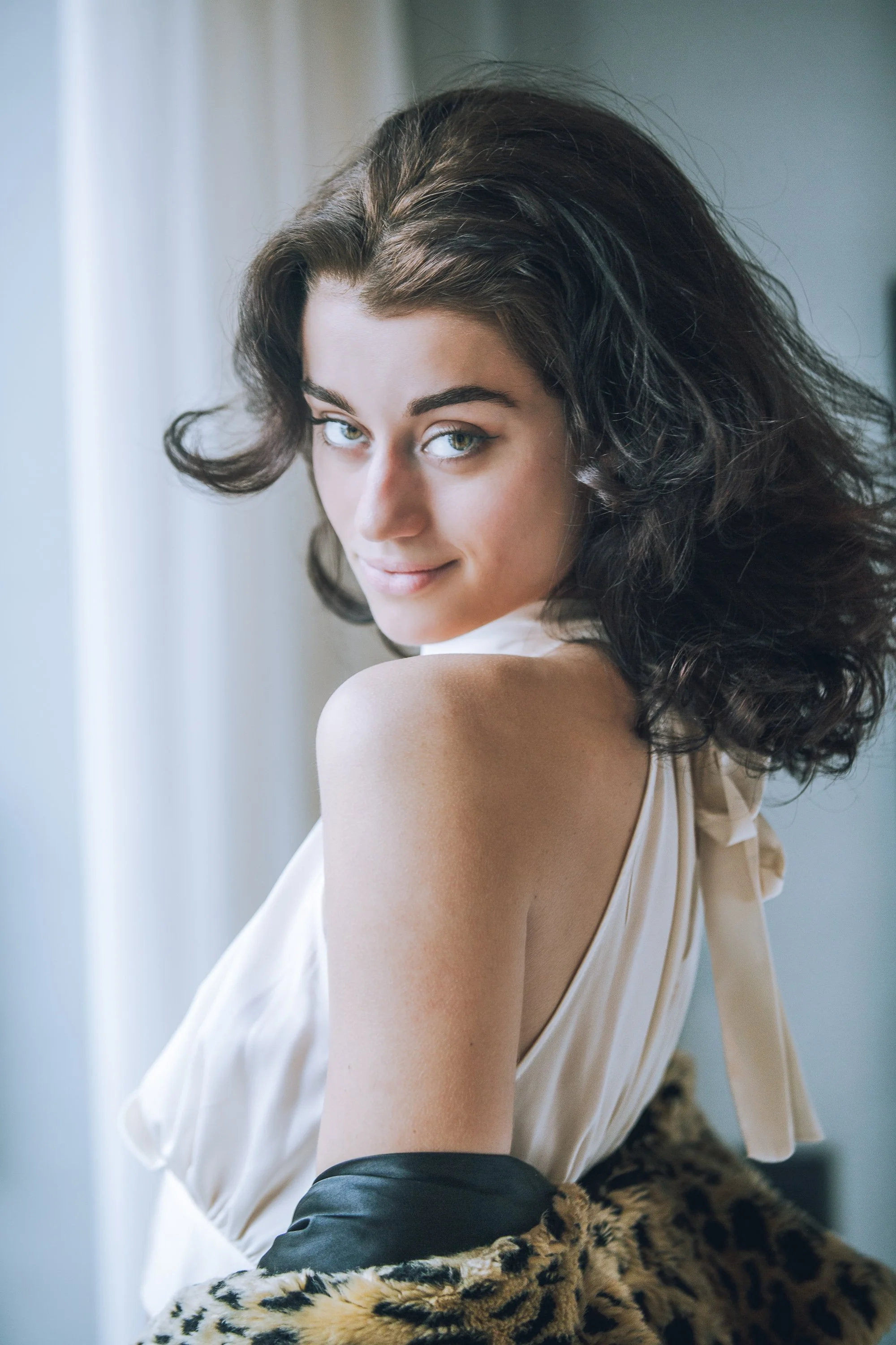 A woman with curly brown hair and gray eyes is sitting near a window, looking at the camera with a slight smile. She is wearing a white satin top with a large bow on the shoulder, a black leather skirt, and is partially covered with a faux fur leopard-print blanket.