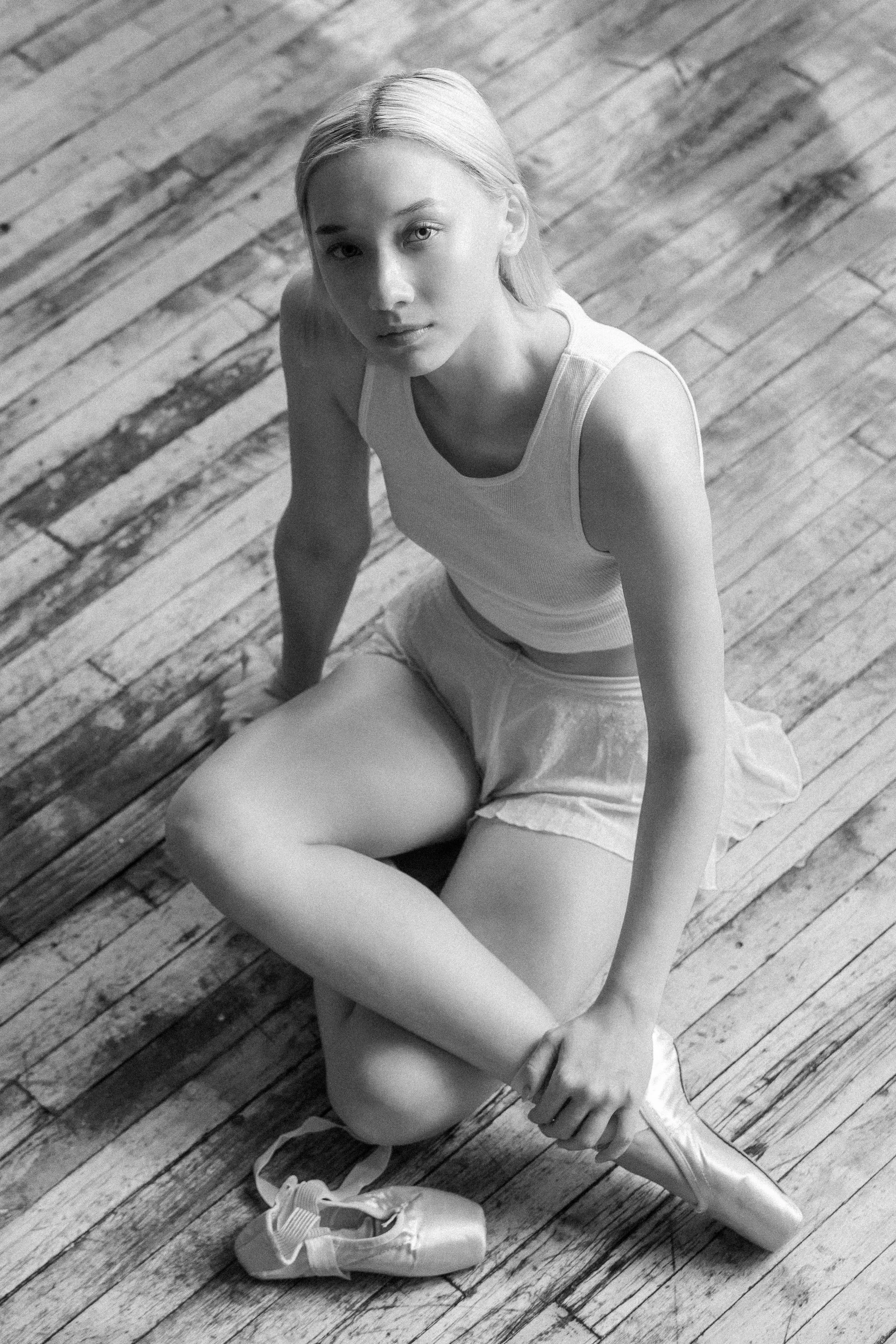 Black and white photo of a young woman with blonde hair sitting on a wooden floor, looking up at the camera with a neutral expression. She is wearing a sleeveless top, shorts, and ballet shoes, with one ballet shoe off and placed on the floor nearby.