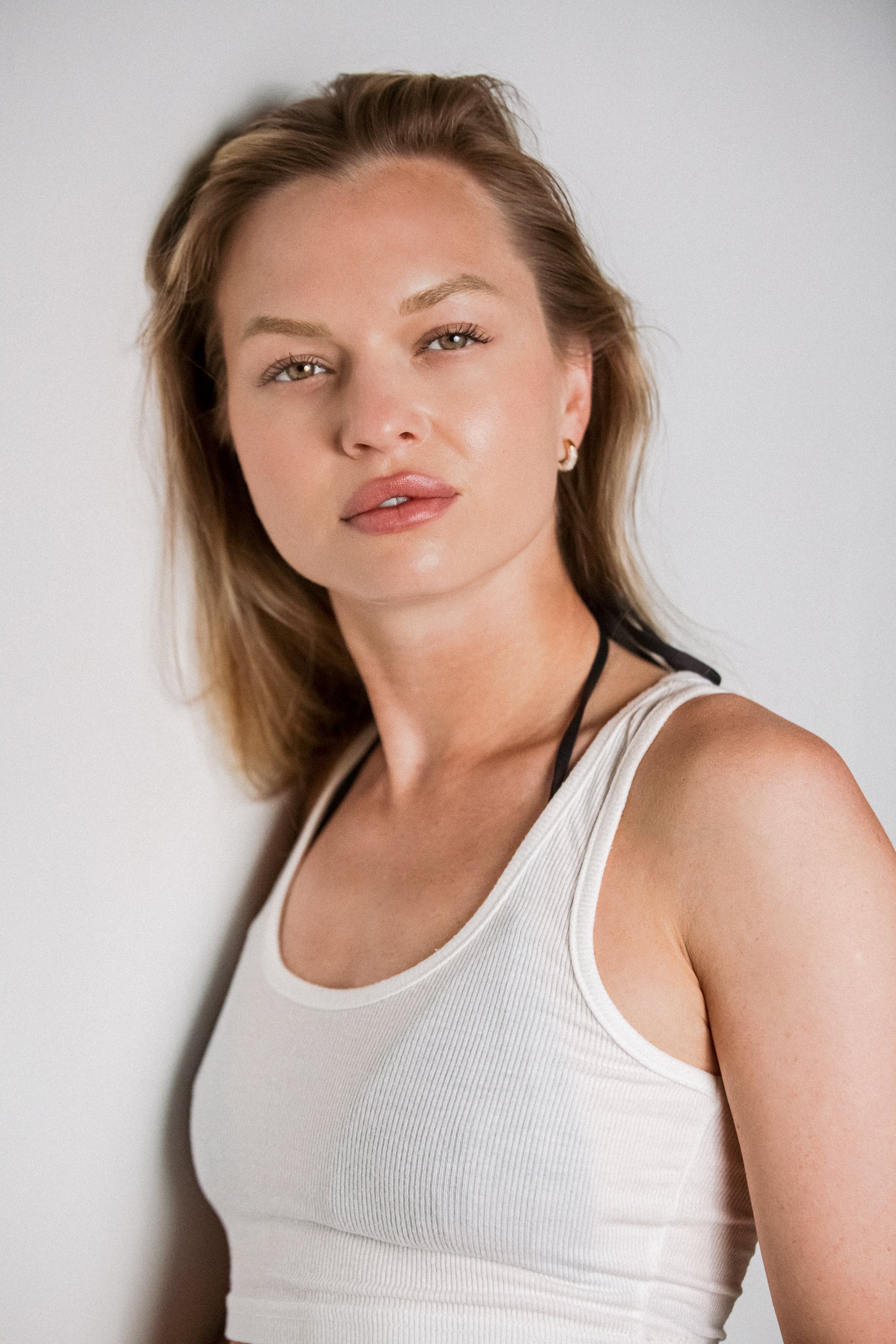 Close-up of a woman with fair skin, light brown hair, and green eyes wearing a white tank top and small gold hoop earrings, standing against a plain light gray background.