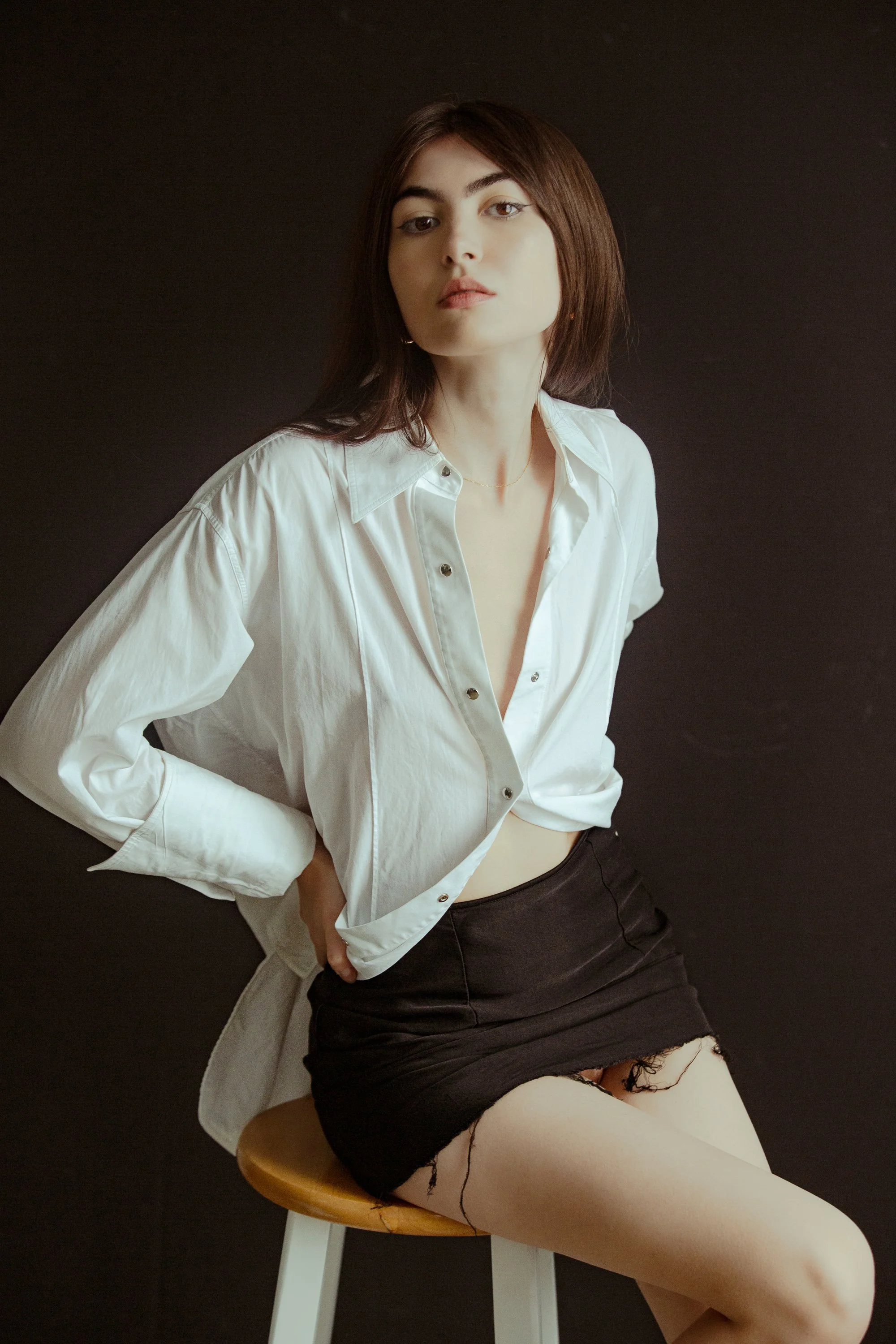 A woman with shoulder-length brown hair wearing a white button-up shirt, black skirt, and white tights with ripped holes, sitting on a wooden stool against a dark background.