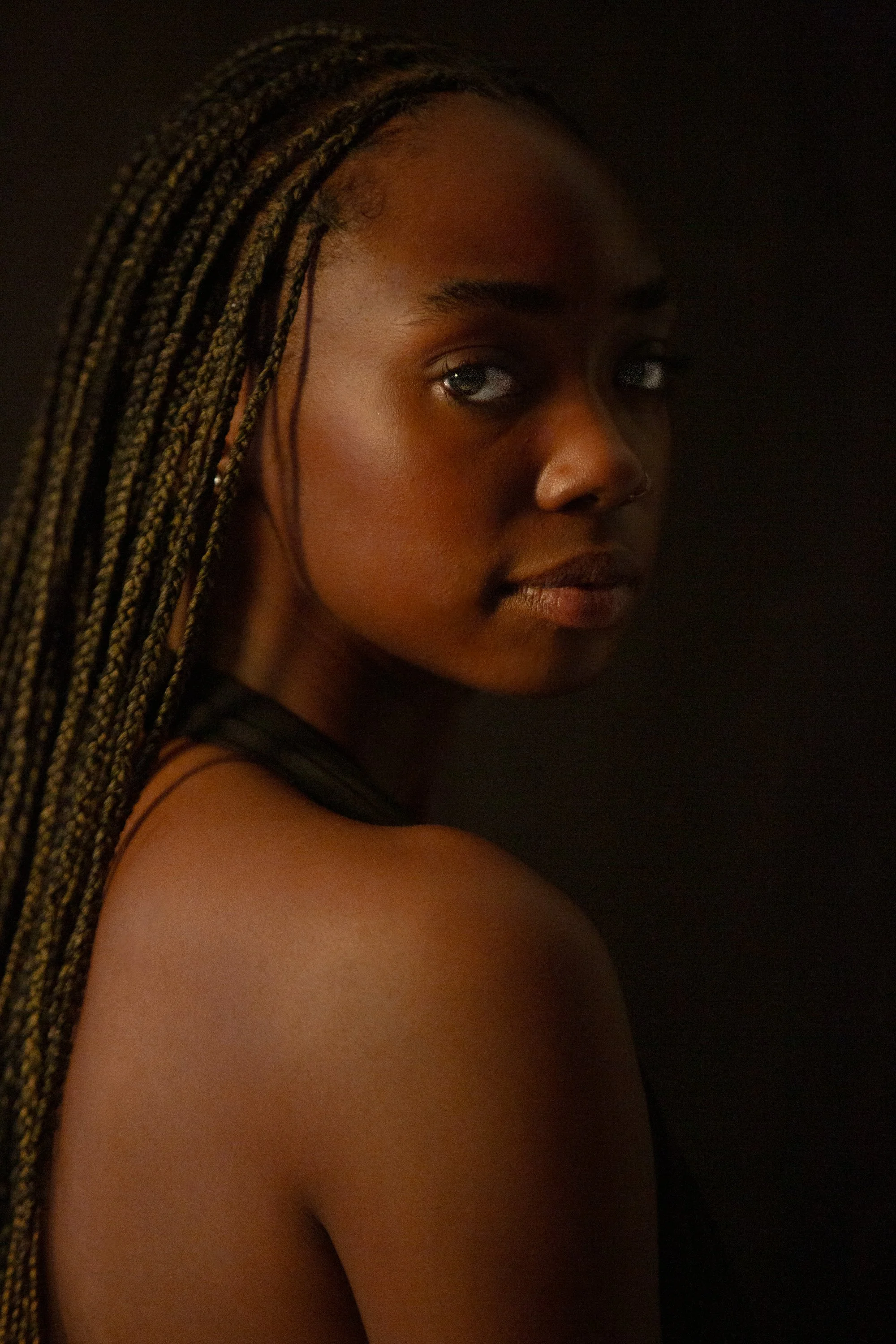 Close-up of a woman with braided hair, dark background, looking over her shoulder.
